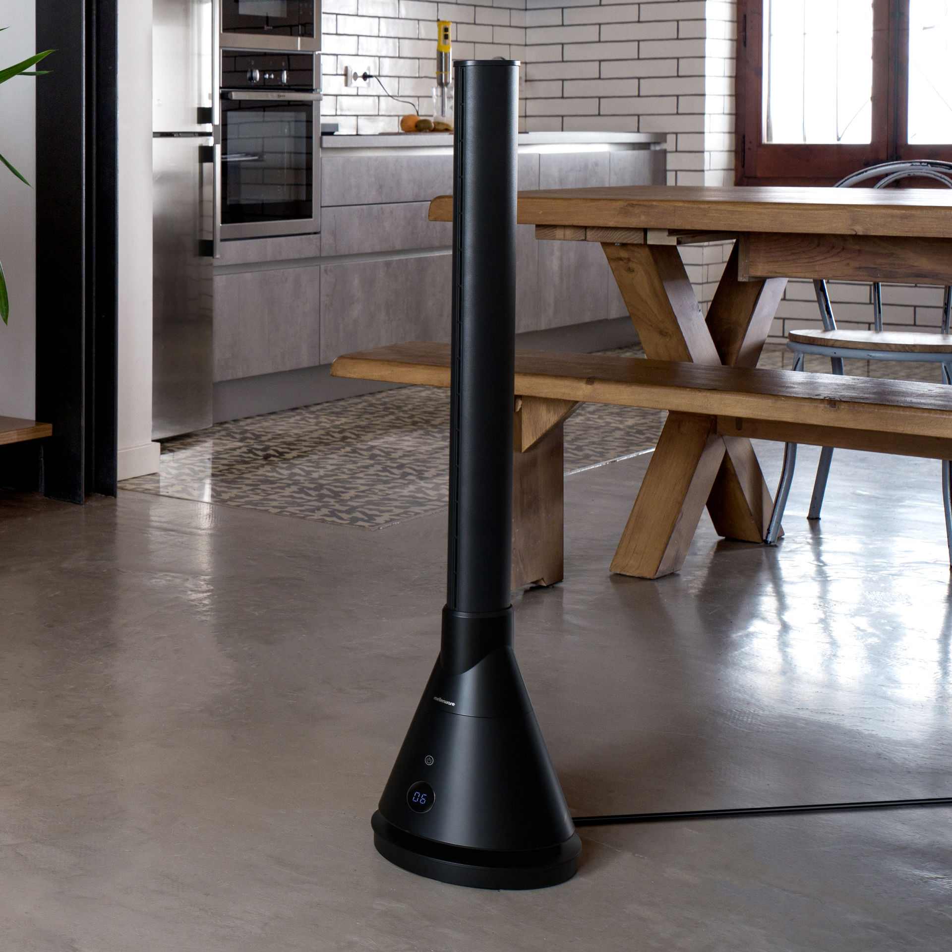 Ventilador de torre negro en una cocina moderna. Fondo: mesa, sillas y electrodomésticos de acero inoxidable.