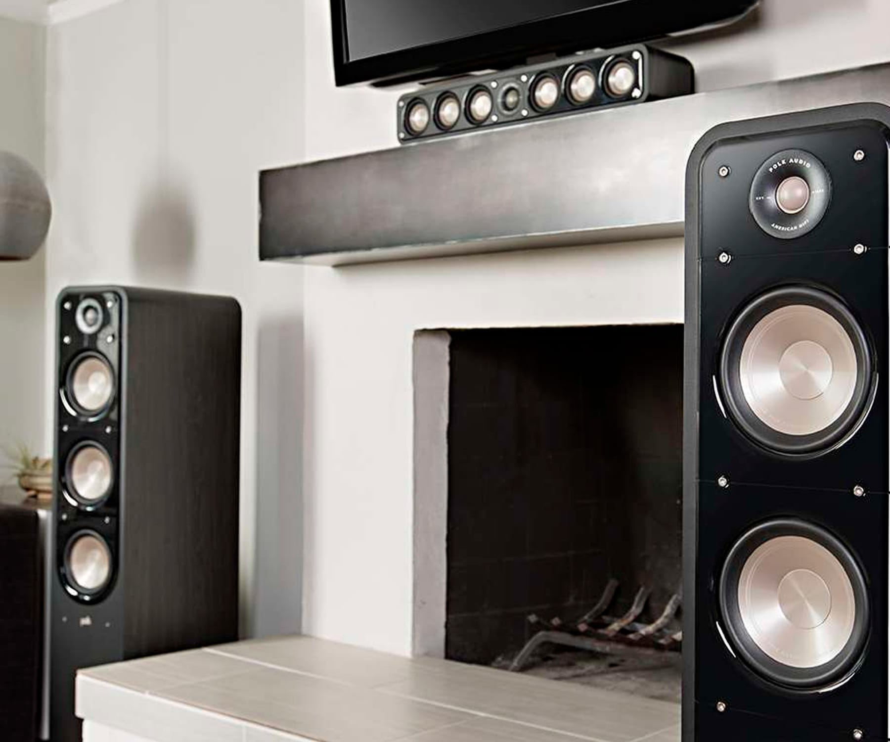 Dos altavoces negros y una barra de sonido encima de una chimenea. Los altavoces están a ambos lados del fuego.