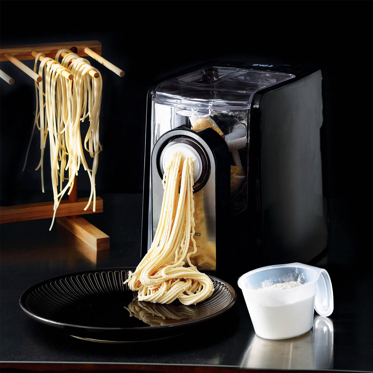 Macchina per pasta che estrae tagliatelle. Tagliatelle appese a un supporto. Piatto nero e tazza con farina.