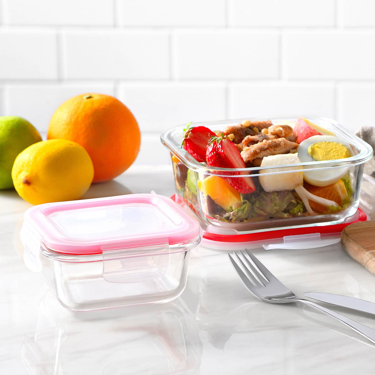 Contenedores de vidrio para alimentos con tapa rosa. Hay comida dentro. Frutas y utensilios están sobre una superficie blanca.