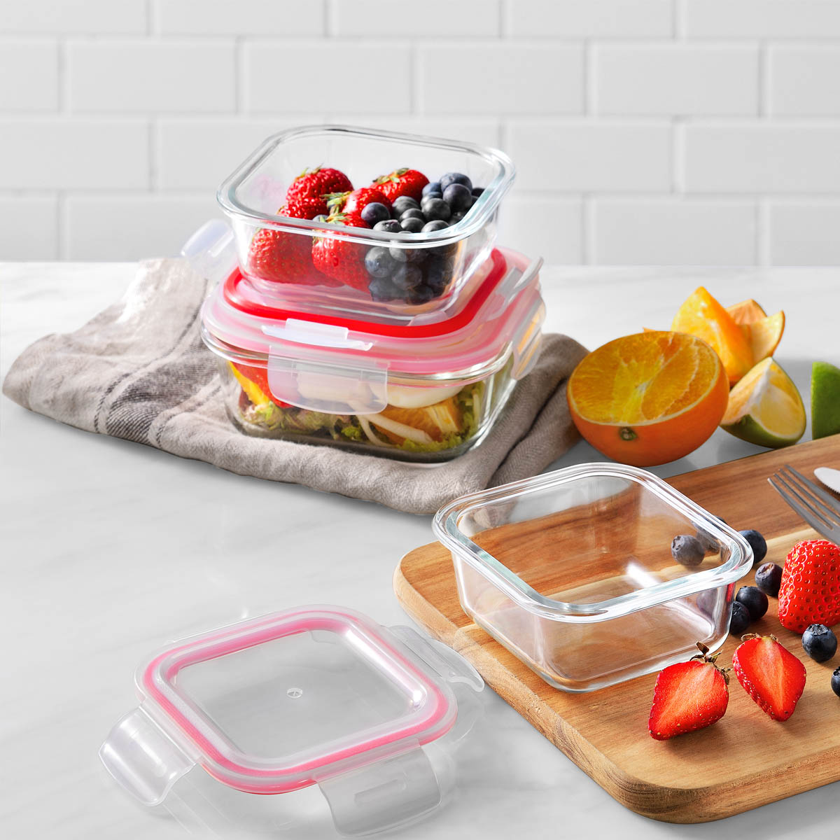 Contenedores de vidrio para alimentos con tapas rojas y frutas, en una tabla de cortar de madera.