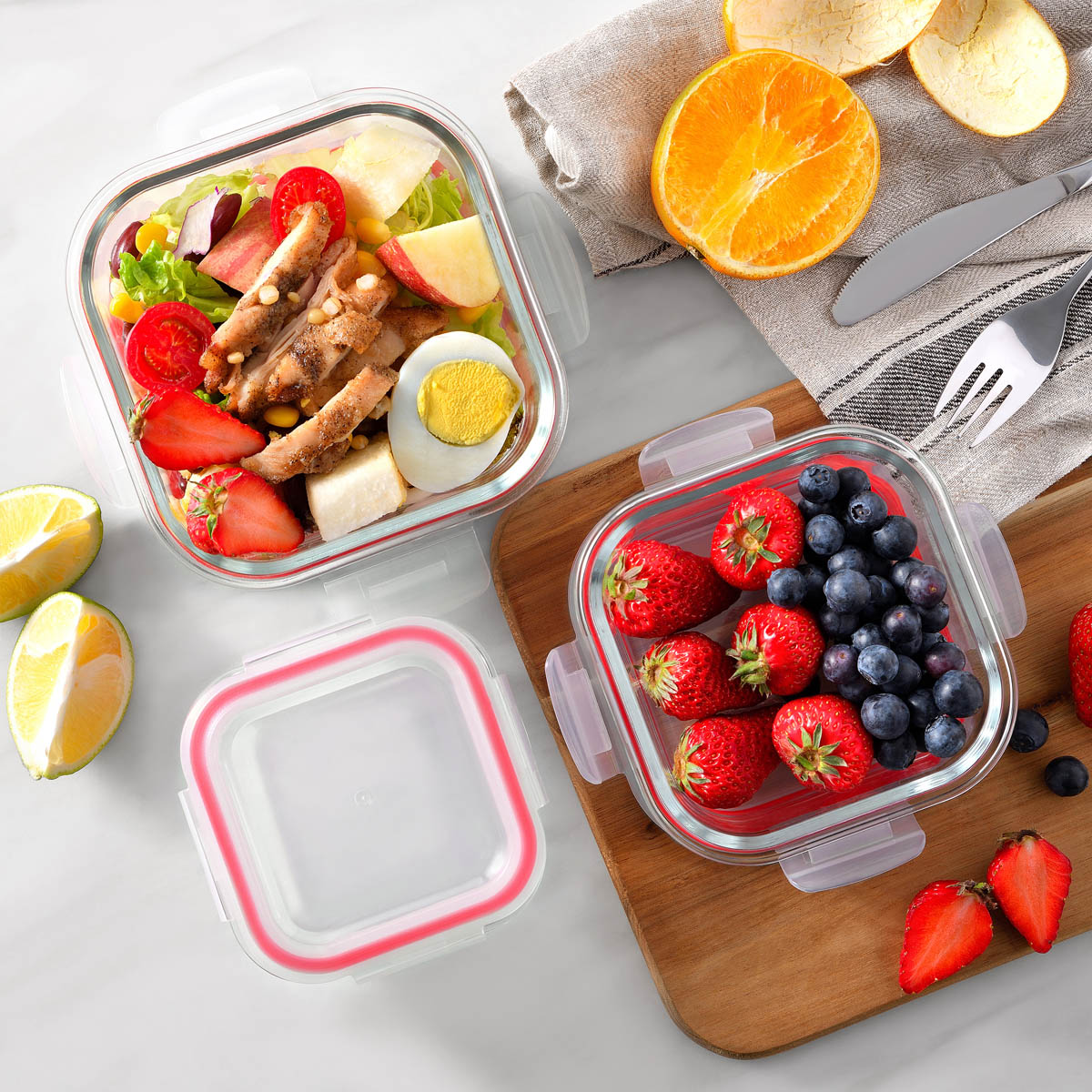 Contenedores de comida de vidrio con ensalada y fruta, con tapas abiertas y cerradas, sobre tabla de madera.