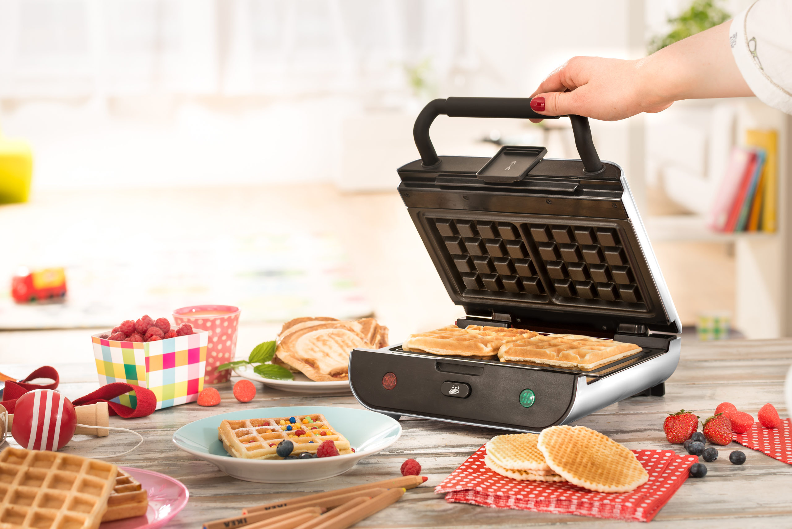 Una persona usa una gofrera, mostrando gofres haciéndose con ingredientes en una mesa. Luz solar y cocina de fondo.