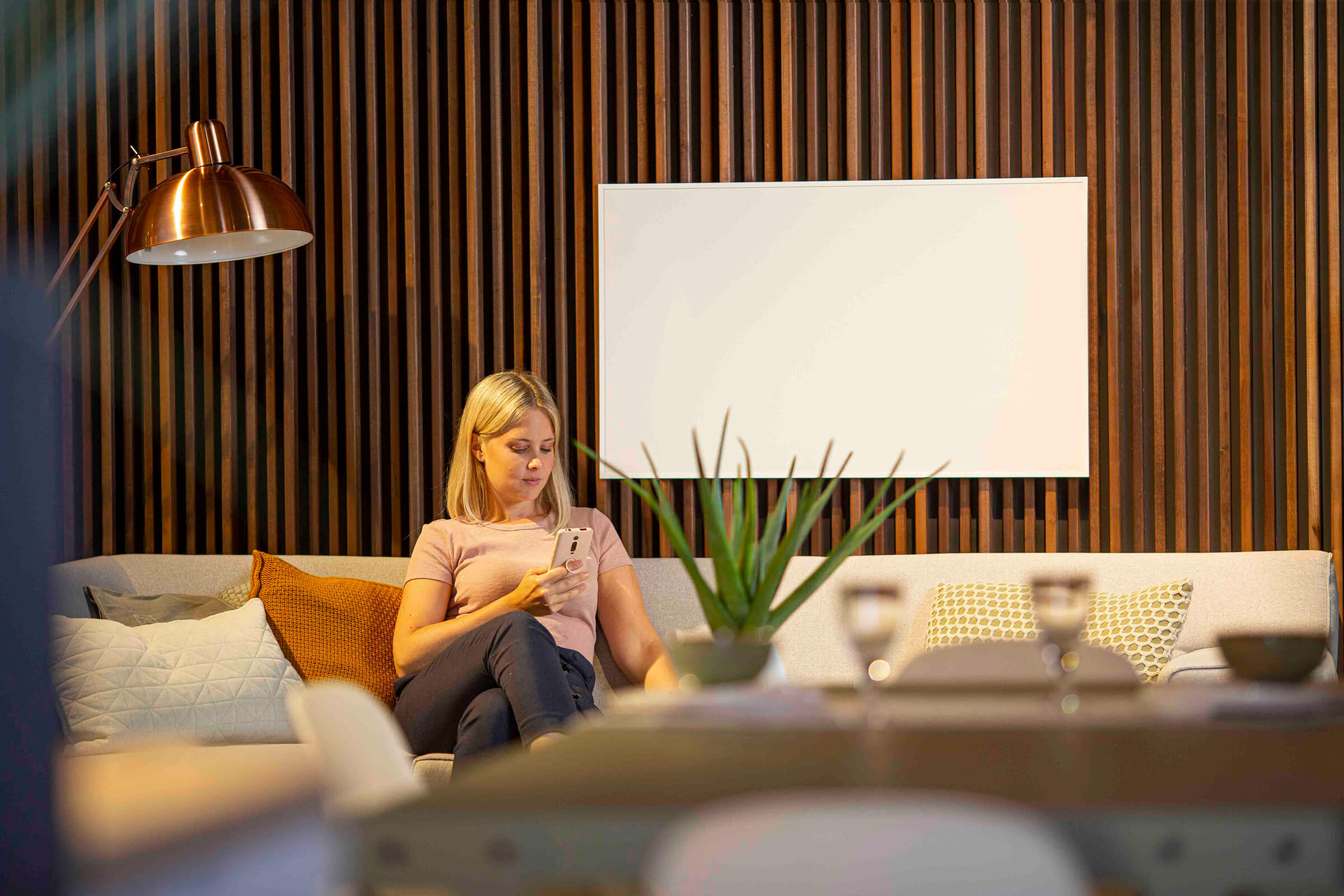 Frau auf Sofa mit Telefon. Holzlattenwand, weiß gerahmtes Kunstwerk und Tisch mit Objekten im Vordergrund.