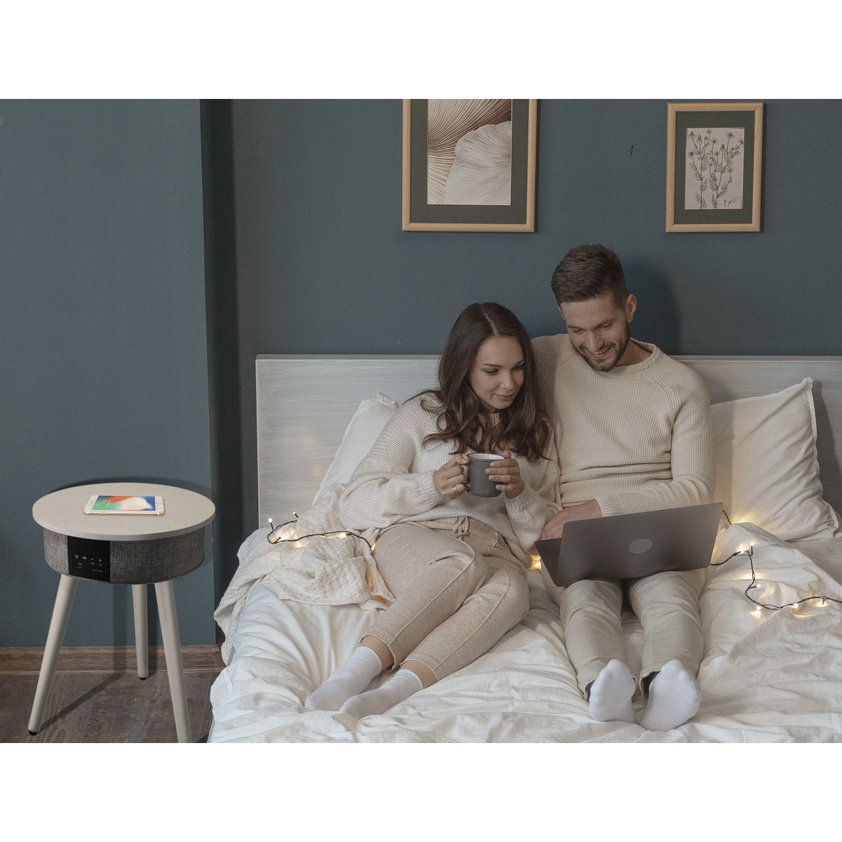 Pareja en la cama con portátil y mesita auxiliar. La mesa tiene un teléfono y un cargador inalámbrico.