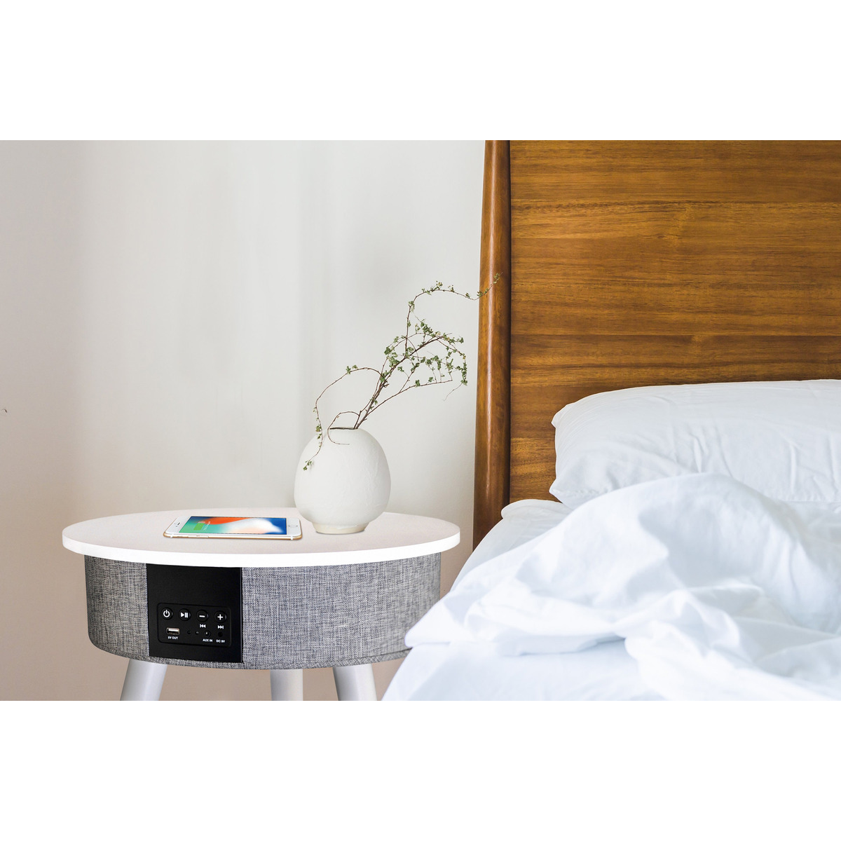 Mesita de noche redonda con teléfono, jarrón y planta. Altavoz abajo. Sábanas blancas en la cama.