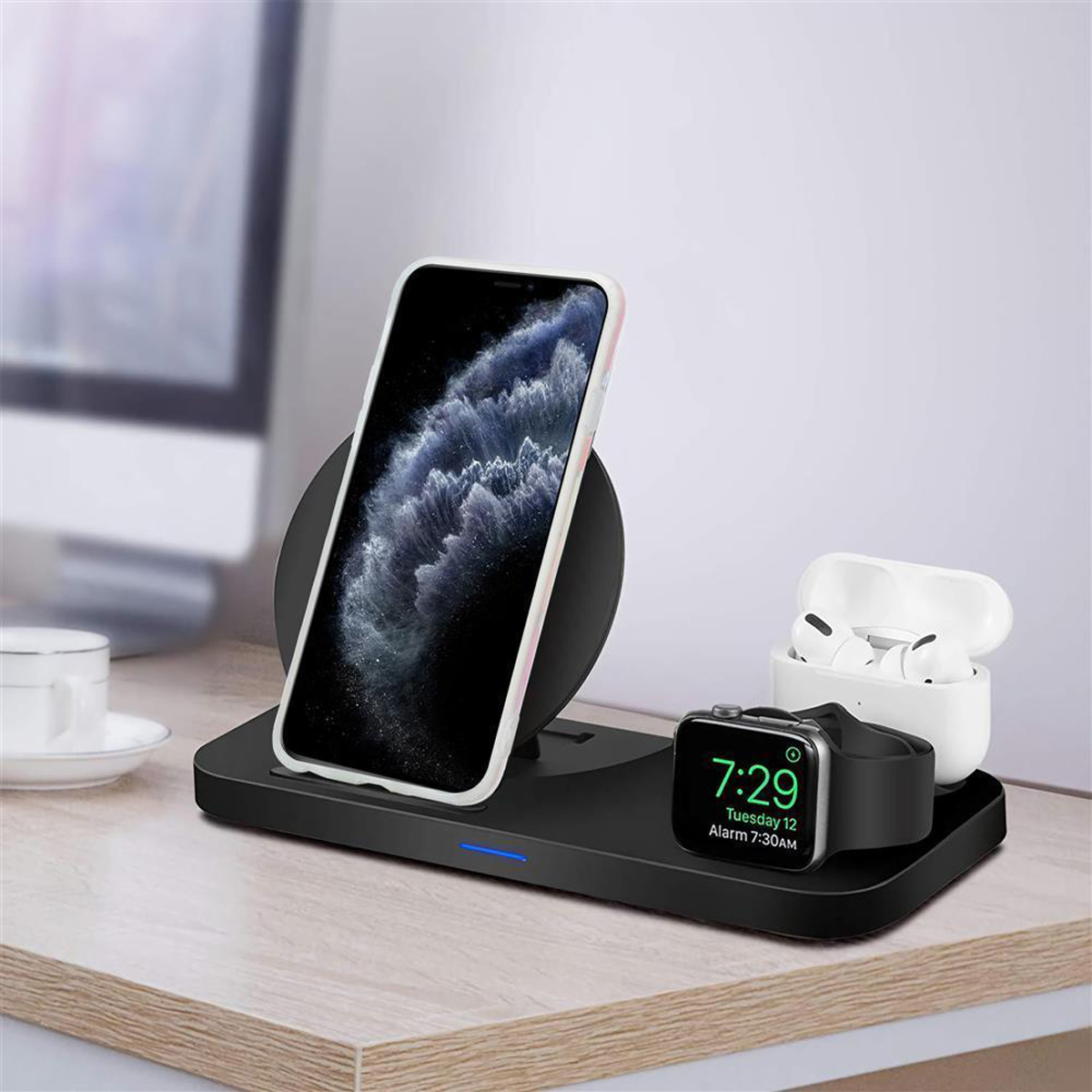 Una stazione di ricarica con telefono, orologio e auricolari su una scrivania. Una tazza bianca e un monitor sullo sfondo.