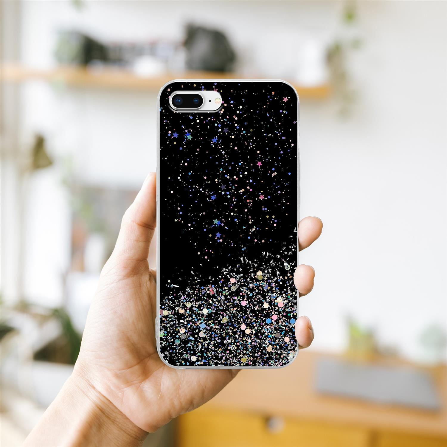 Hand hält eine Handyhülle mit Glitzer. Schwarzer Hintergrund mit Glitzer und Sternen. Heller Innenhintergrund.