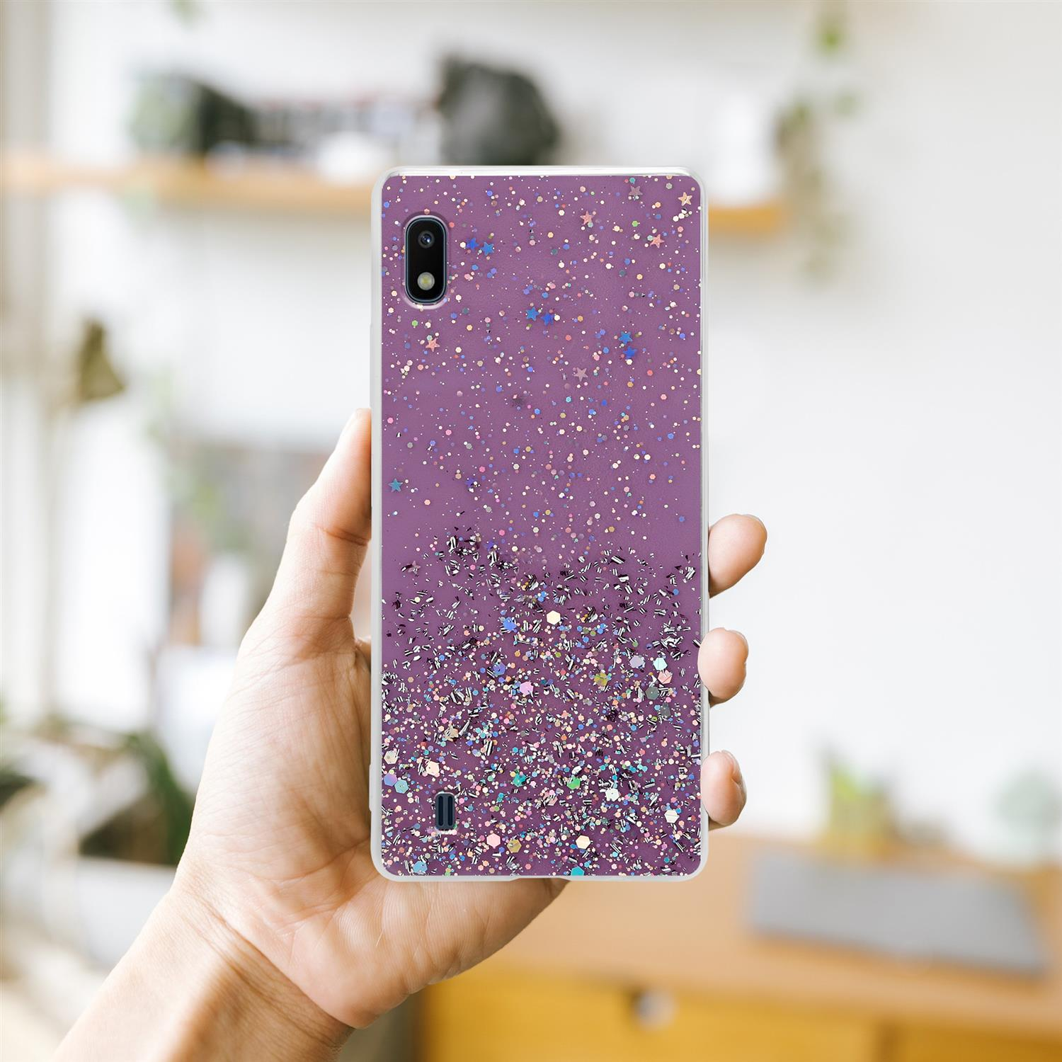 Eine Hand, die ein Telefon mit einer lila glitzernden Hülle hält. Der Hintergrund ist verschwommen und zeigt einen Raum.