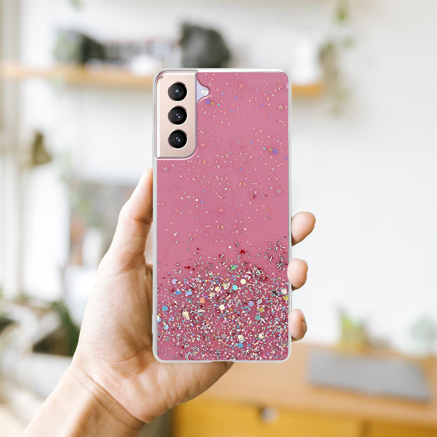 Una mano tiene un telefono con una custodia rosa ricoperta di glitter. Lo sfondo è una stanza sfocata.