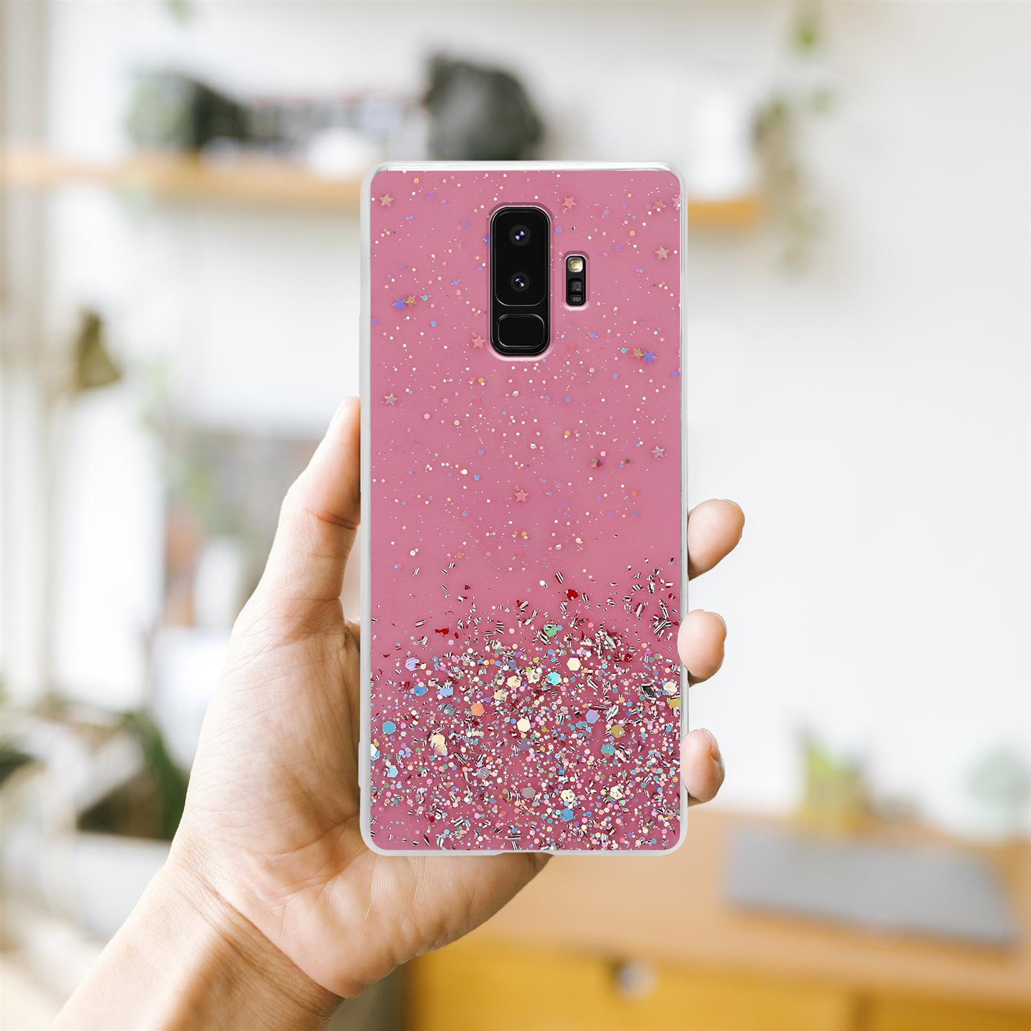 Hand hält eine rosa Handyhülle mit Glitzer. Hintergrund ist ein verschwommener Raum.