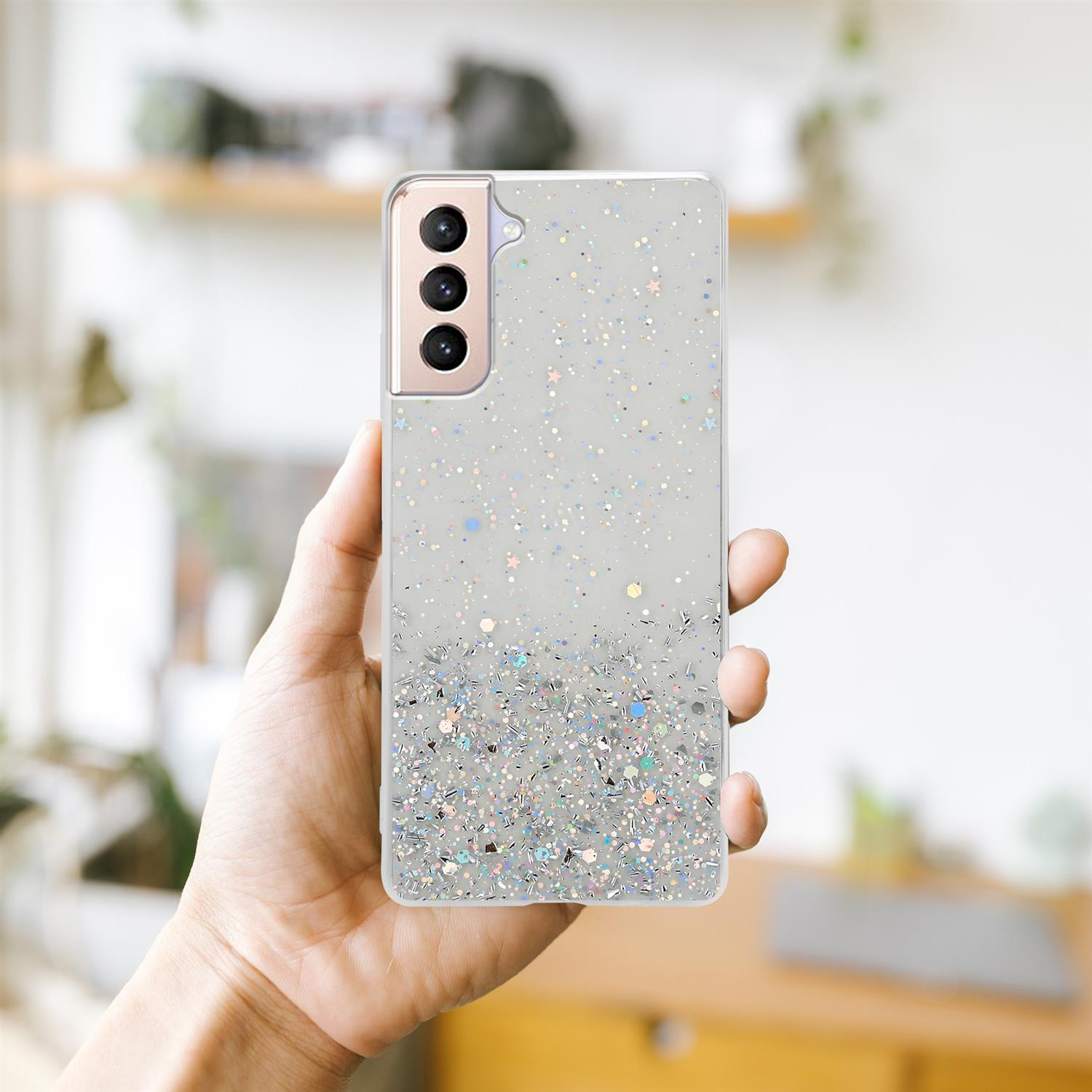 Eine Hand hält ein Telefon mit durchsichtiger Hülle und silbernem Glitzerdesign. Der Hintergrund ist unscharf.