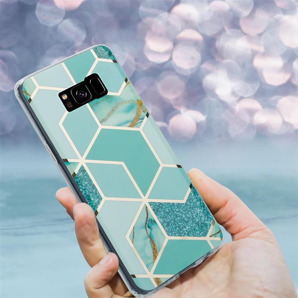 Mano che tiene un telefono. Il telefono ha una custodia. La custodia ha un design geometrico verde e oro. Sfondo blu.