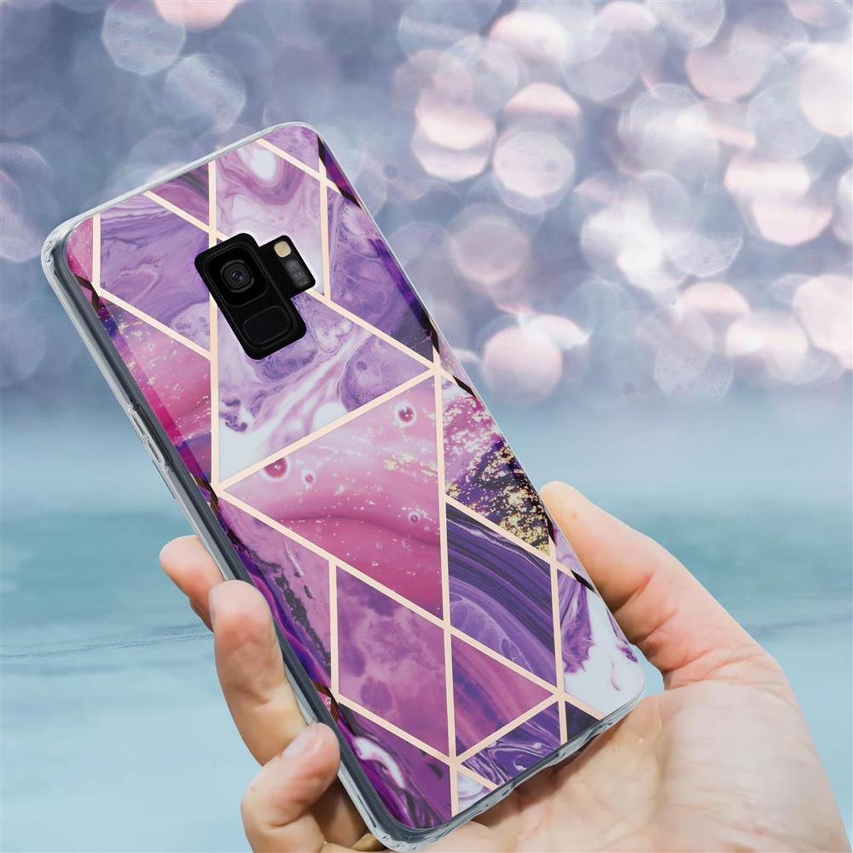 Una mano tiene un telefono con una custodia dal design in marmo viola e oro. Lo sfondo è sfocato.