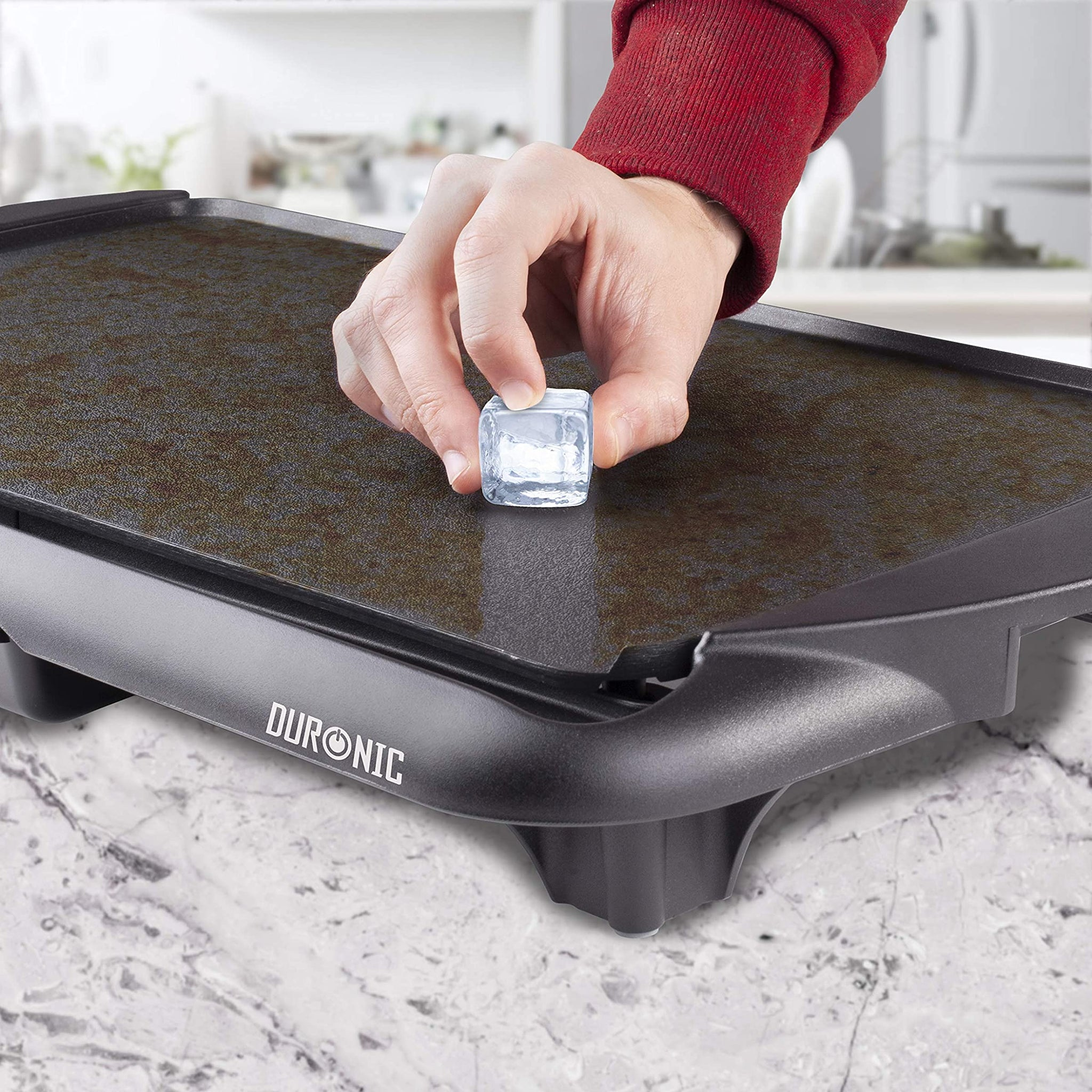 Eine Hand legt einen Eiswürfel auf eine schwarze elektrische Grillplatte, DUONIC-Markenname sichtbar.