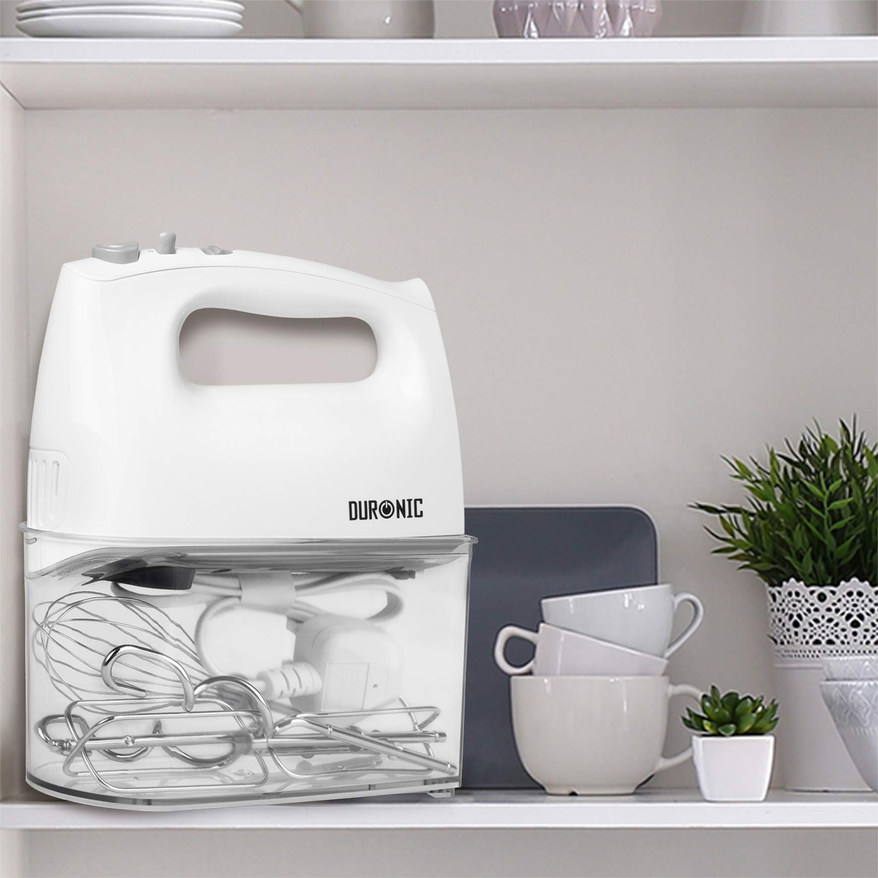 Weißer Mixer mit Zubehör in einem transparenten Behälter. Der Behälter befindet sich in einem Schrank auf einem Regal.
