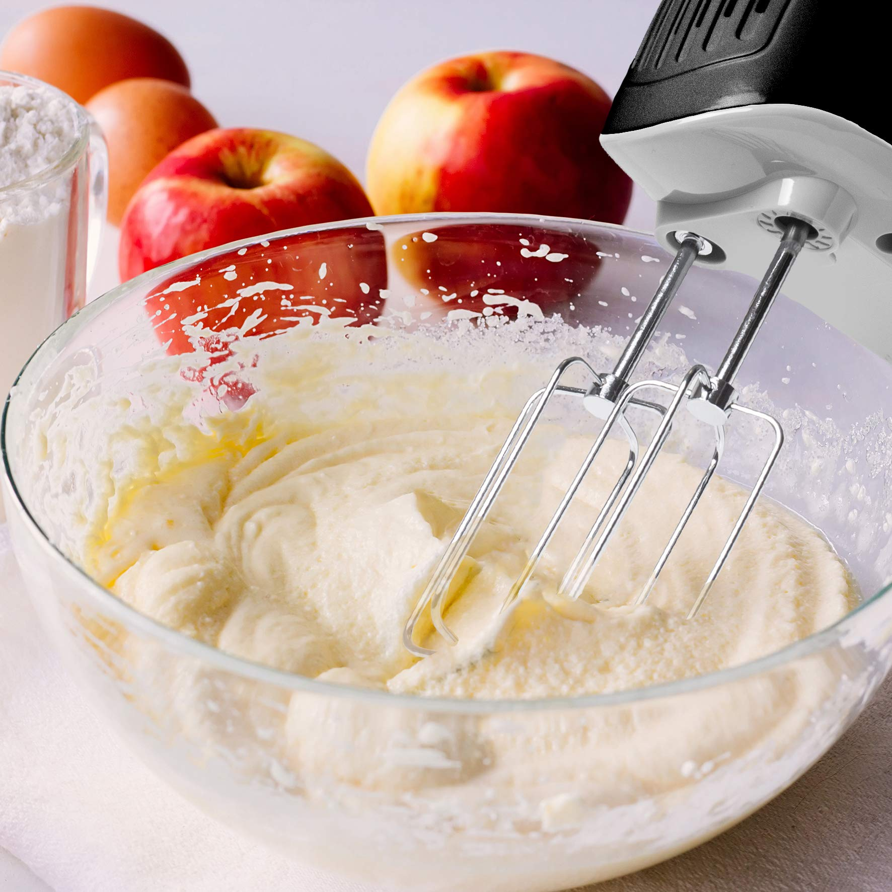 Un bol de vidrio con masa blanca siendo mezclada con una batidora de mano. Manzanas y huevos al fondo.