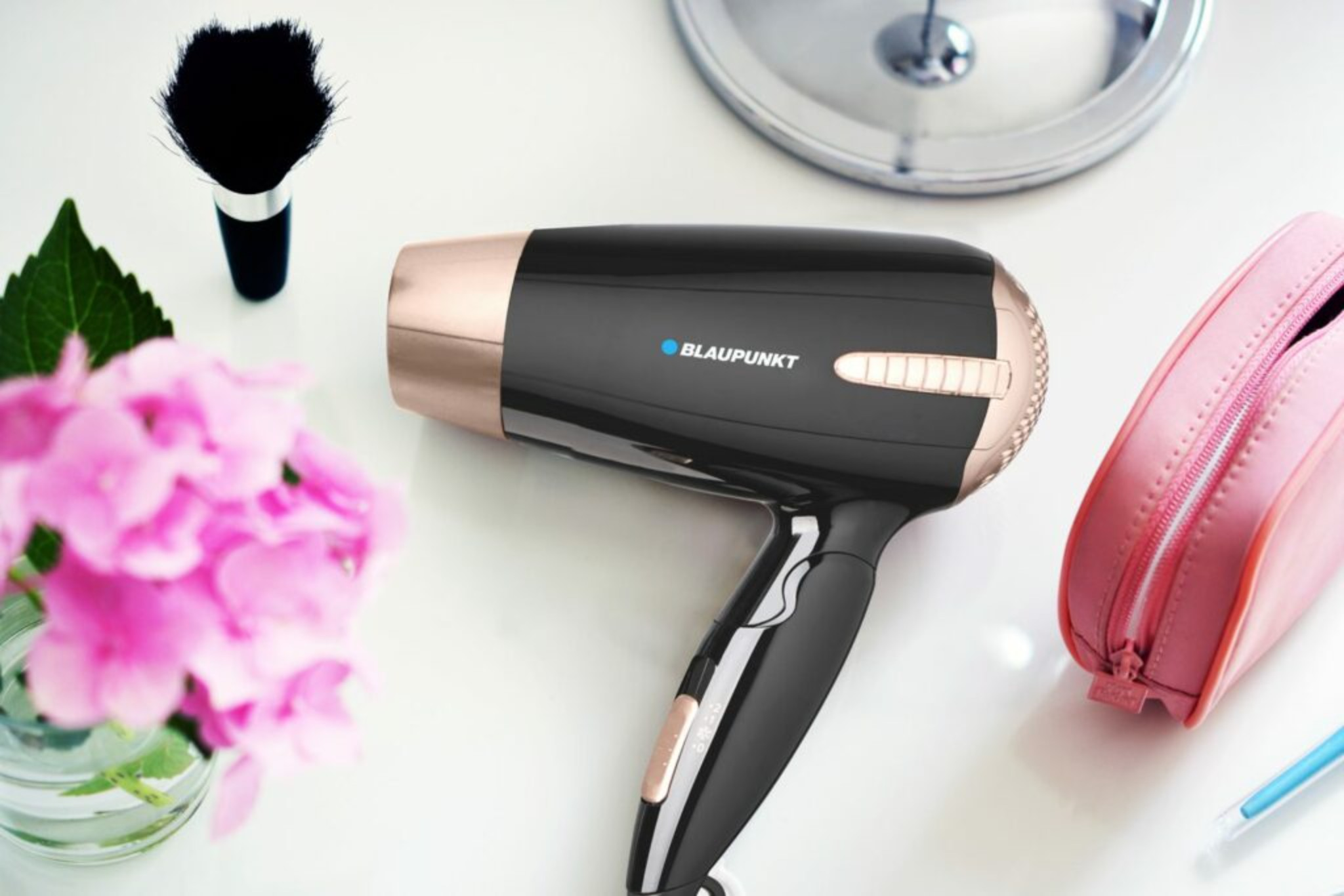 Secador de pelo negro y oro rosa, brocha de maquillaje, estuche rosa y espejo sobre superficie blanca.