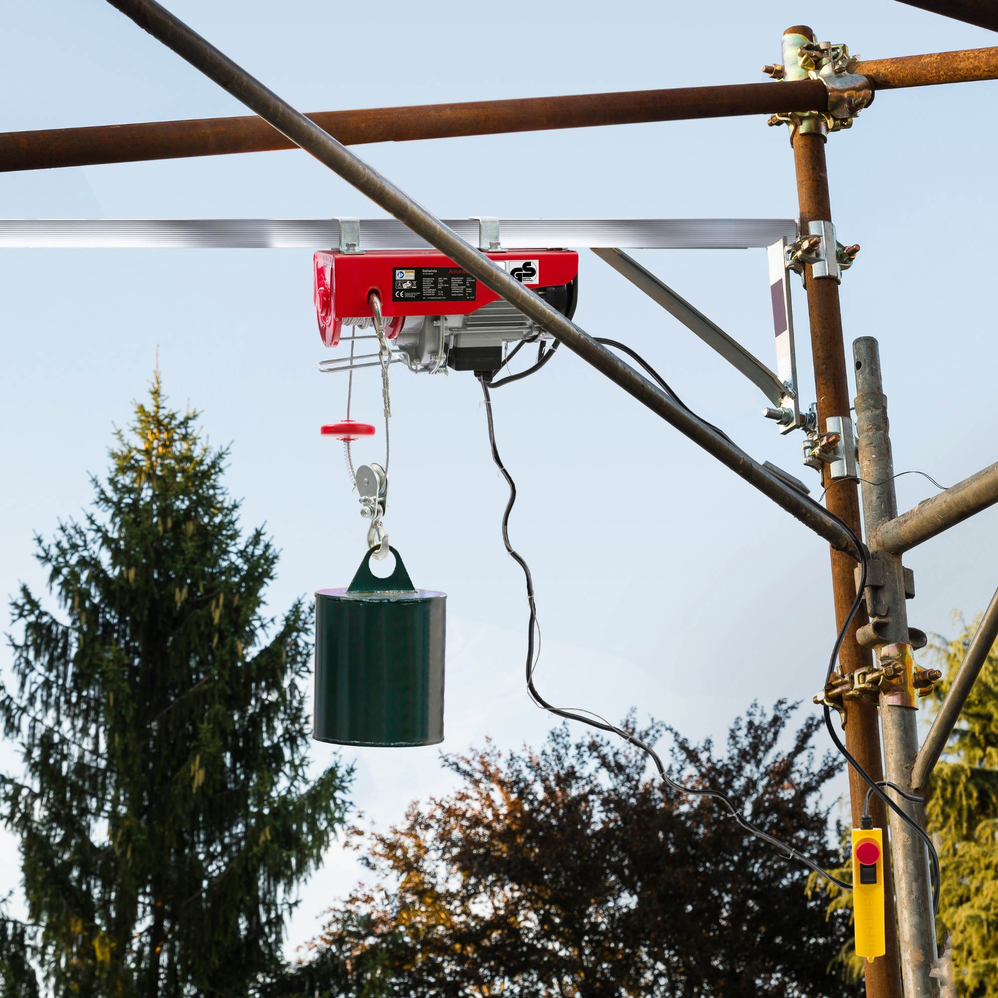 Ein roter Elektro-Hebezug hängt an einem Gerüst und hält ein grünes Gewicht. Bäume und Himmel im Hintergrund.