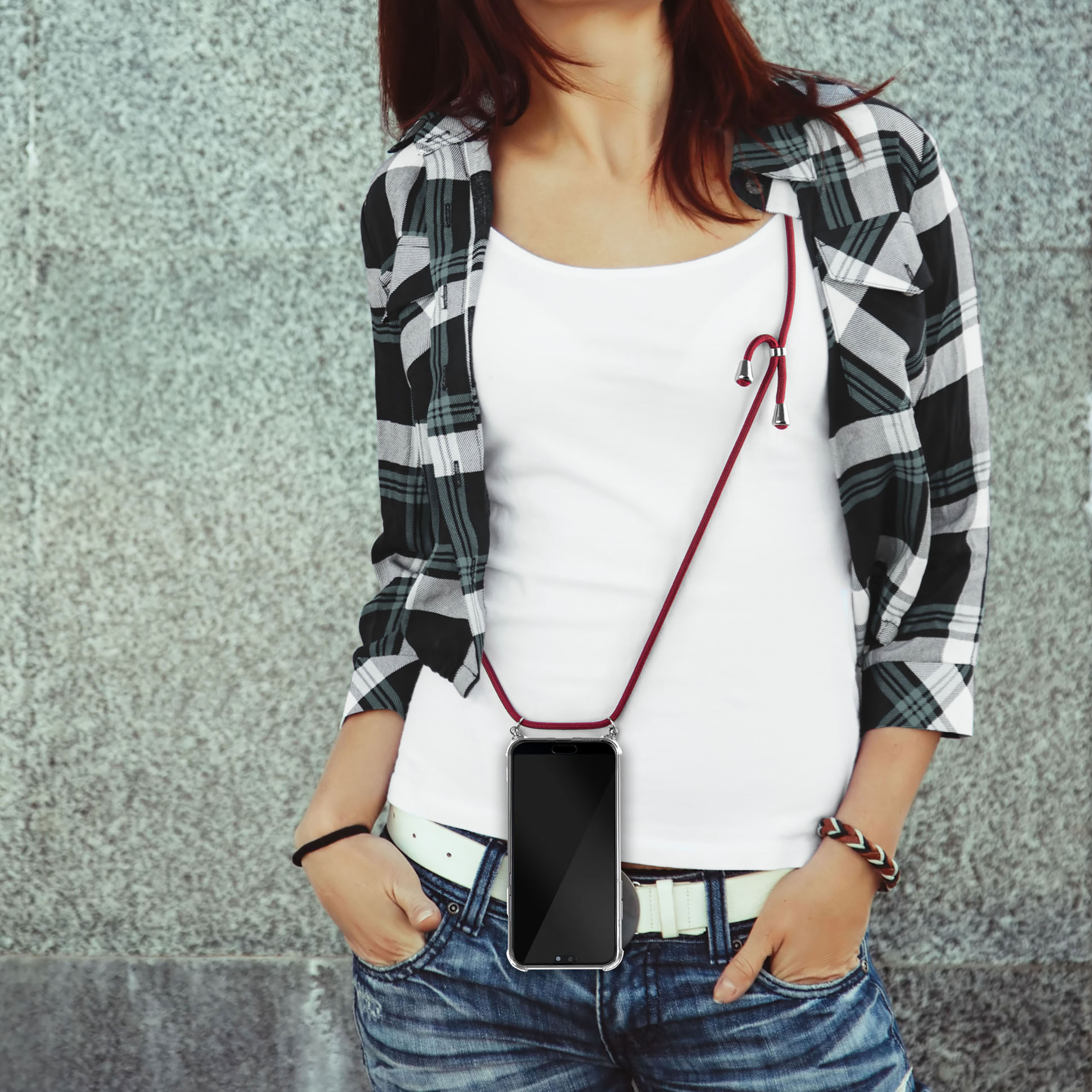 Eine Frau mit kariertem Hemd, weißem Hemd und Jeans hält ein Telefon mit rotem Riemen.