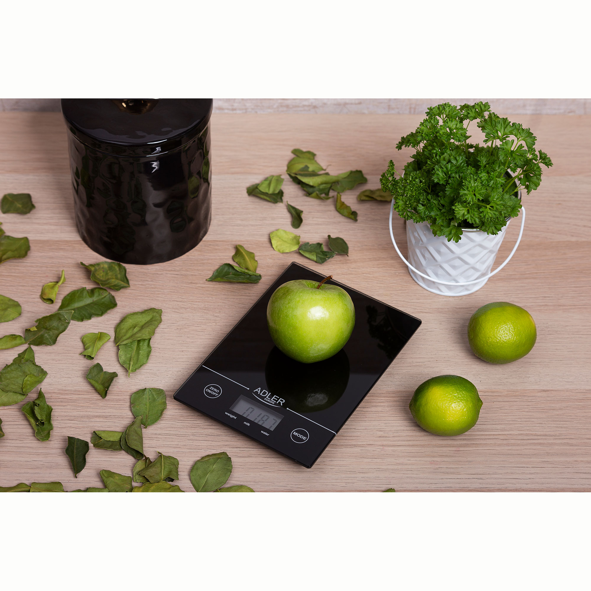 Czarna waga z jabłkiem i limonkami na drewnianej powierzchni. Jest słoik i pietruszka.