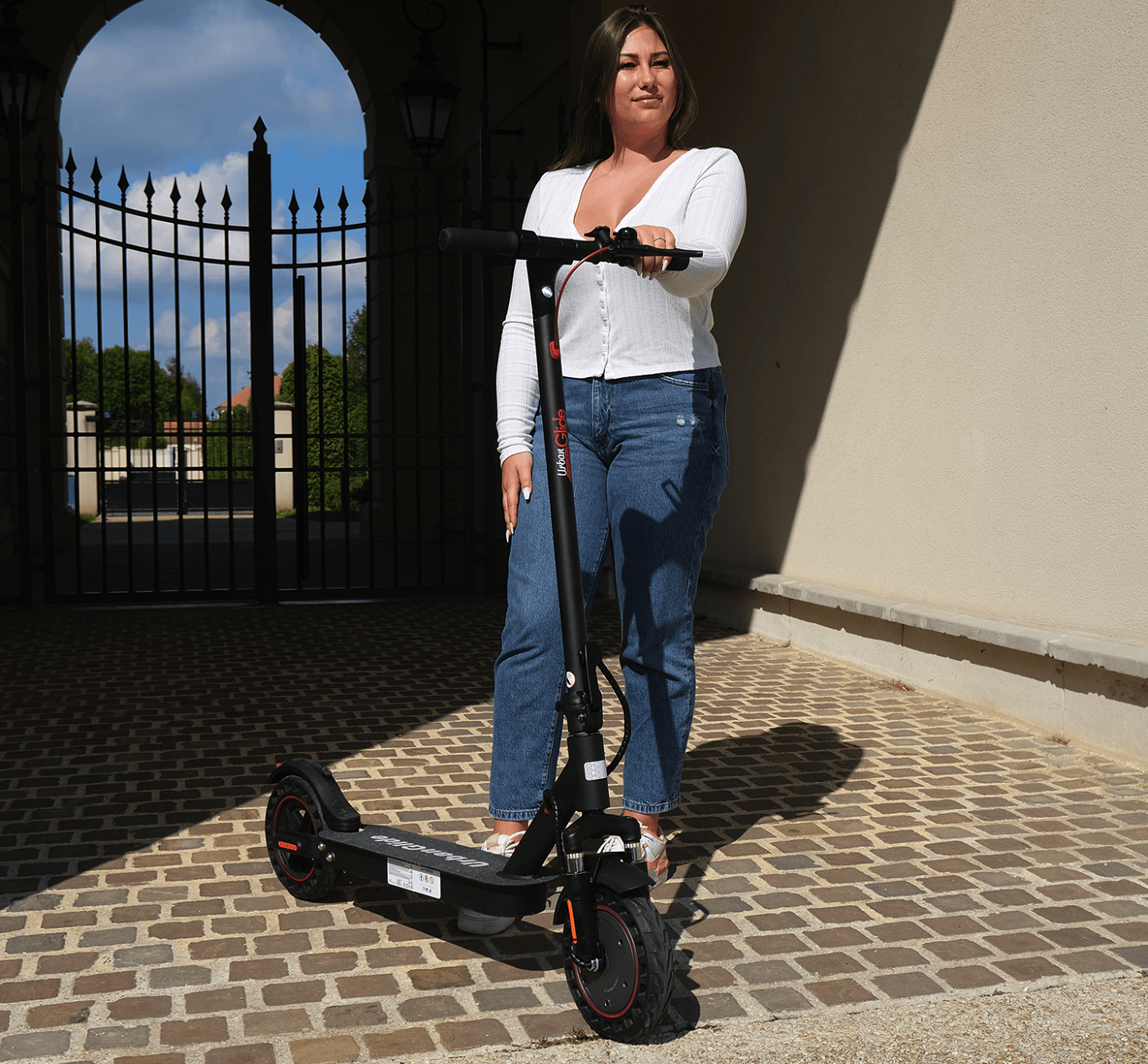 Een vrouw staat met een elektrische scooter voor een gebouw met een poort.