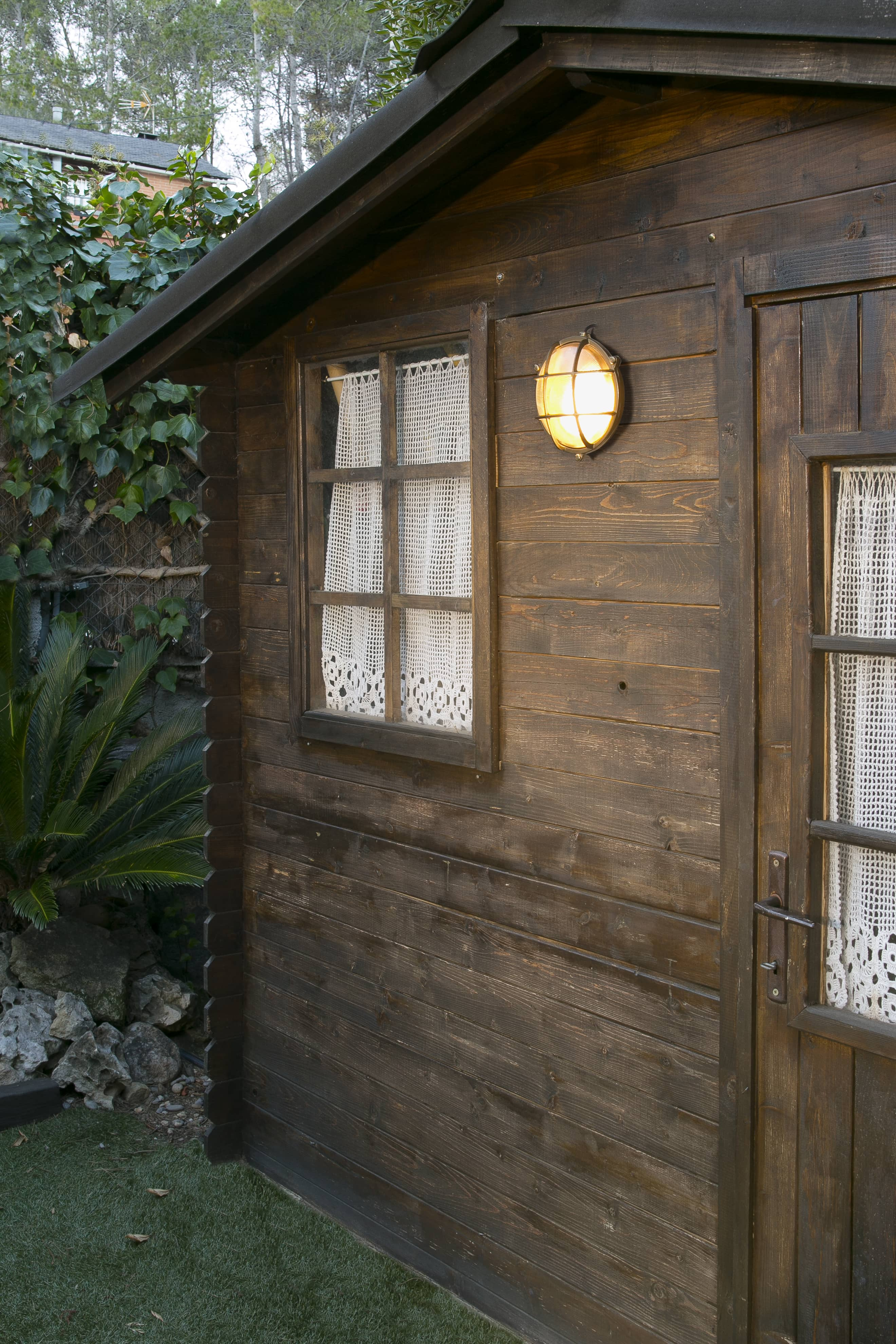 Ein kleiner Holzschuppen mit Fenster und Tür, einer Leuchte und Grün drumherum.