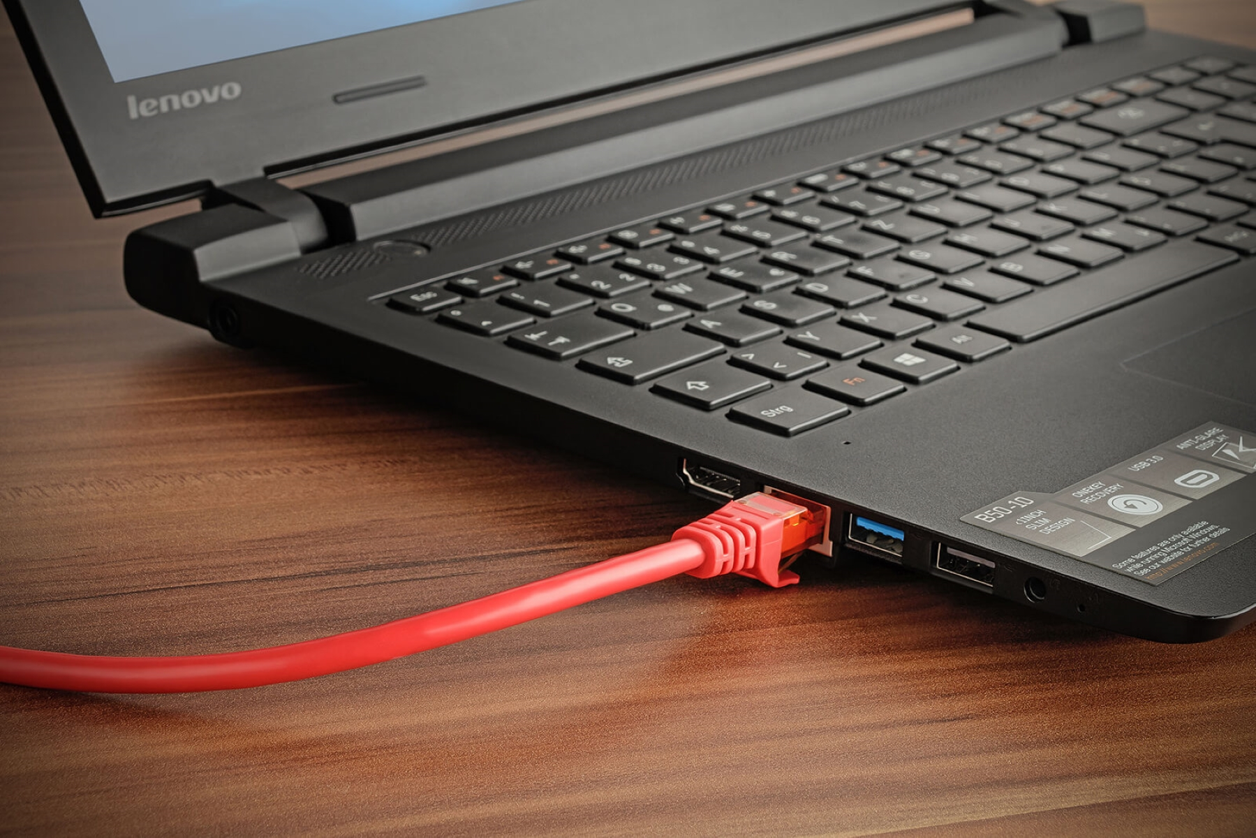 Ein schwarzer Laptop mit einem roten Ethernet-Kabel auf einem hölzernen Schreibtisch.