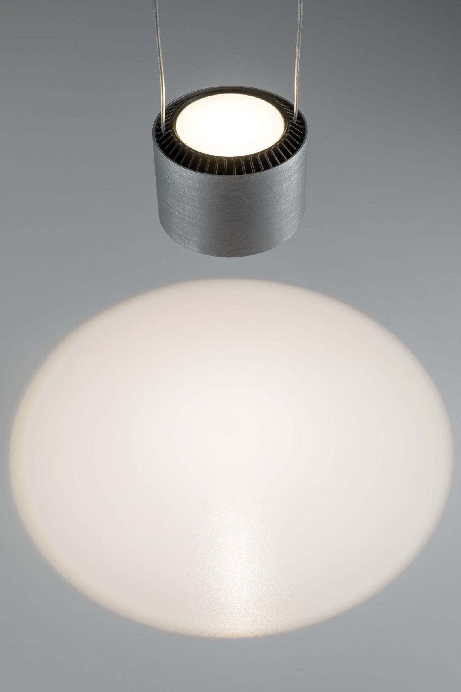 Moderne silberne LED-Pendelleuchte, wirft kreisförmigen Lichtstrahl auf grauen Hintergrund.