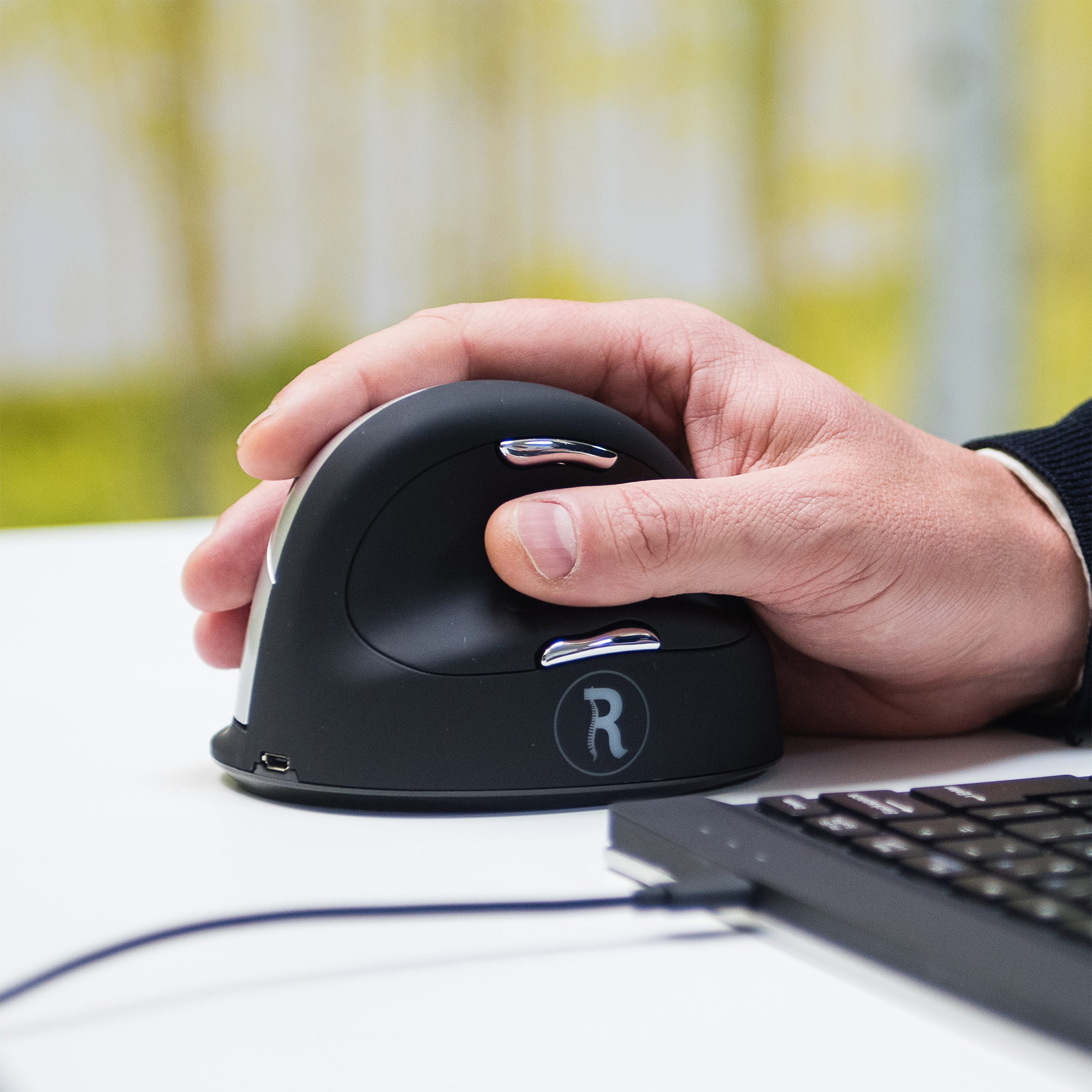 Eine Hand hält eine schwarze ergonomische Maus. Rechts befindet sich eine Tastatur.