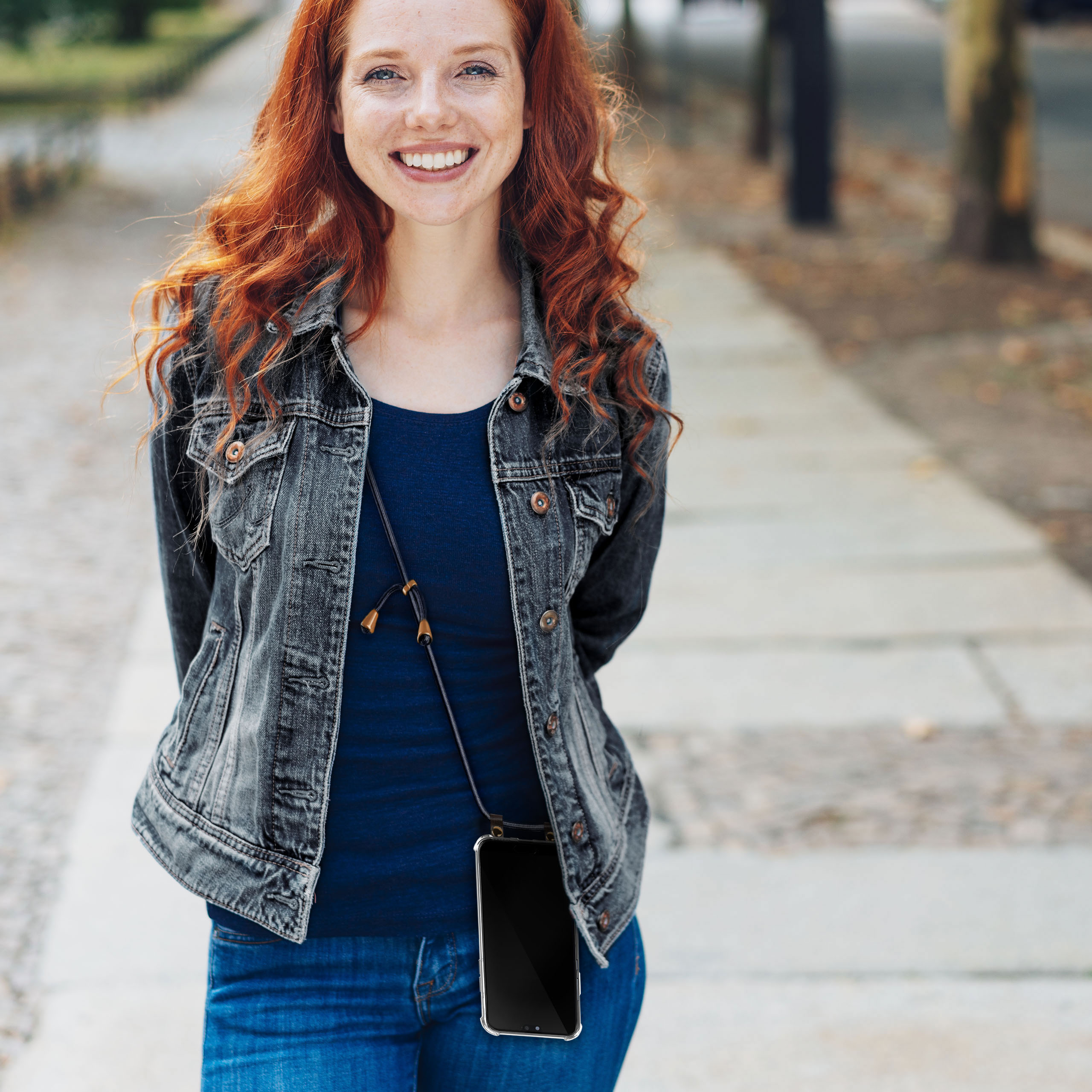 Frau mit roten Haaren lächelt, trägt Jeansjacke und Jeans und hält ein Telefon an einem Gurt.