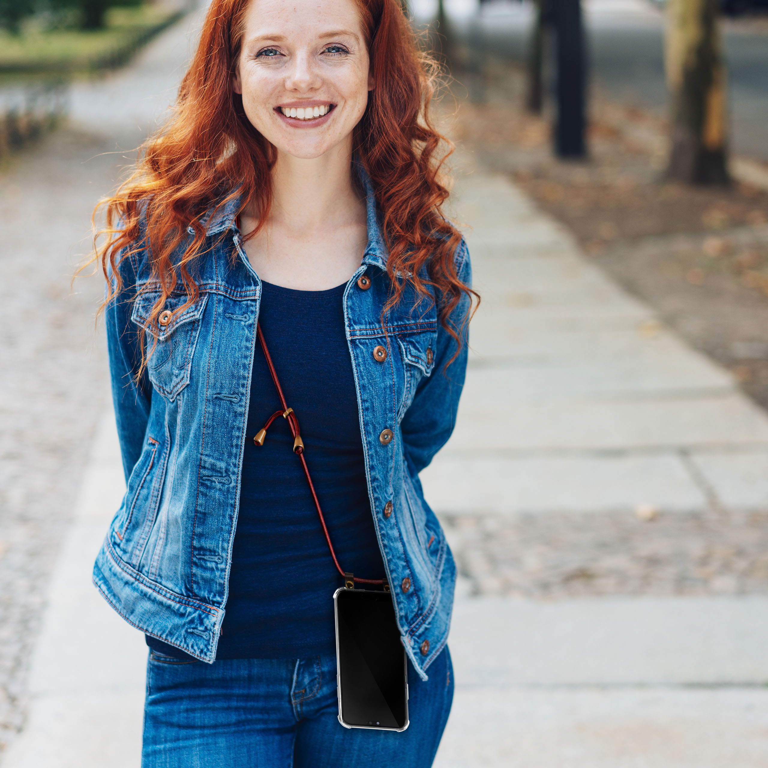 Frau mit roten Haaren, trägt eine Jeansjacke und Jeans, lächelt und hält ein Telefon mit Riemen.