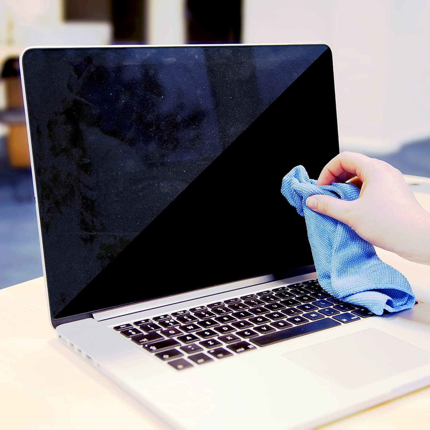 Eine Person reinigt einen Laptop-Bildschirm mit einem blauen Mikrofasertuch auf einem Holzschreibtisch.