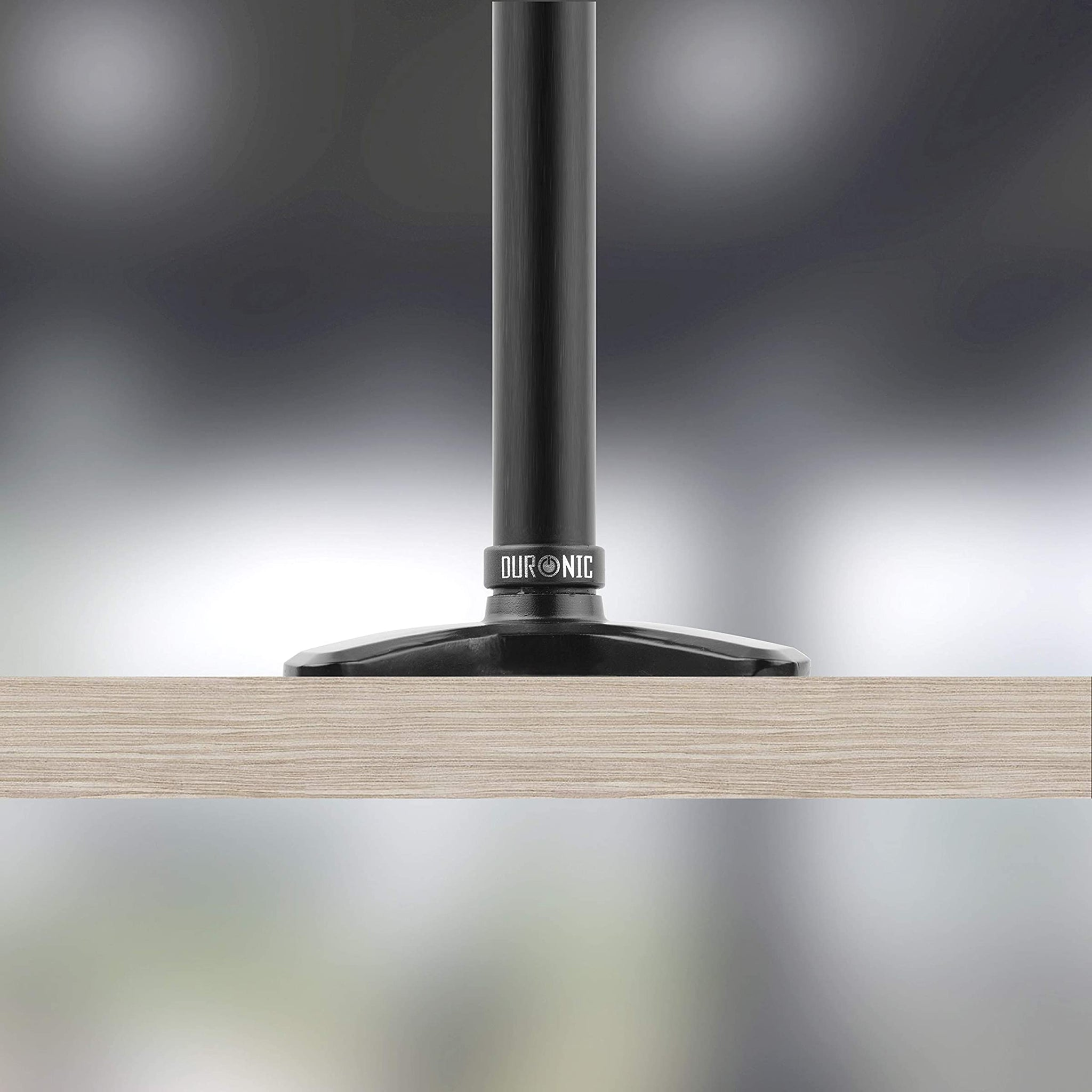 Base de soporte de monitor negro fijada a una mesa de madera, fondo borroso.