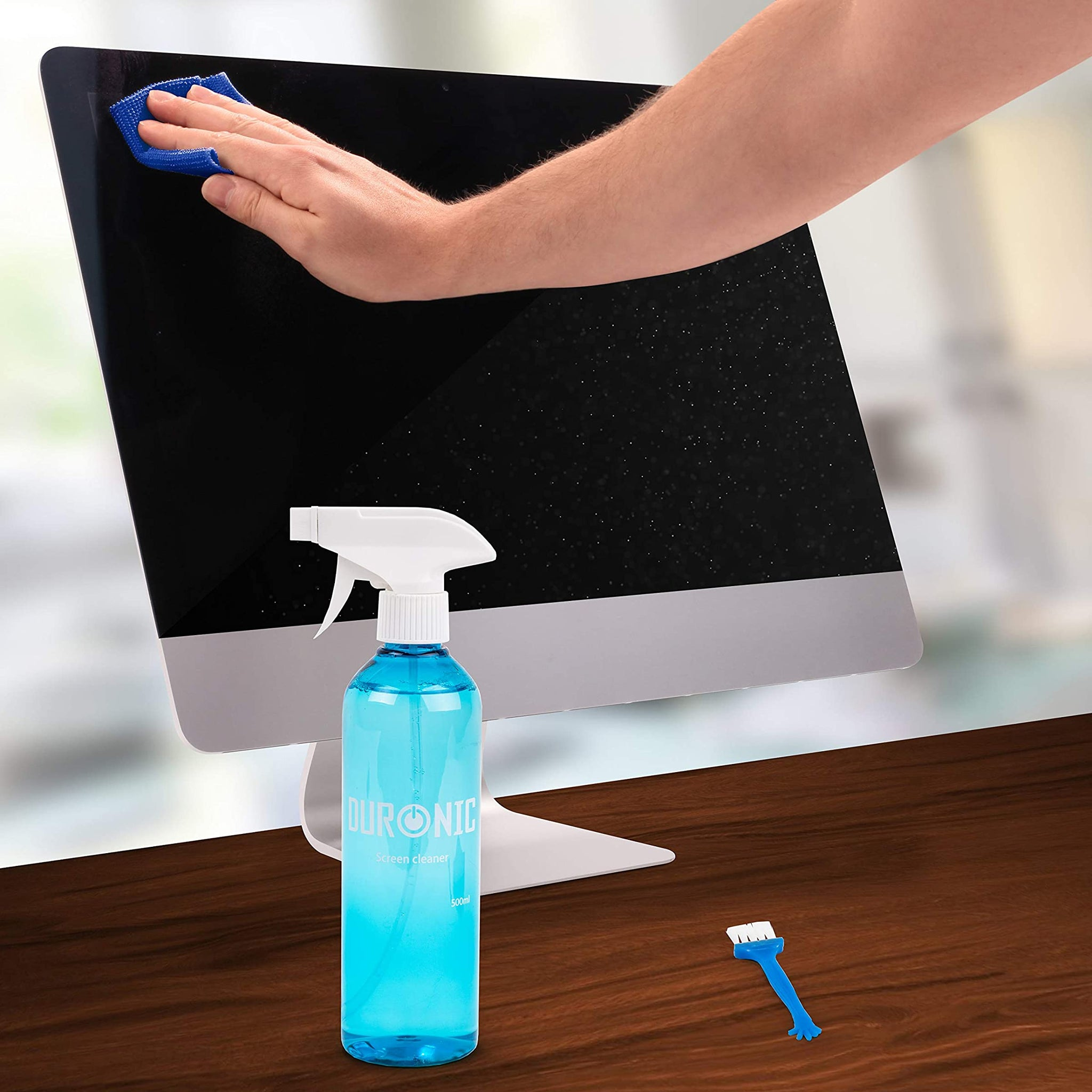 Hand sprüht Bildschirmreiniger auf einen Computermonitor mit einem blauen Mikrofasertuch und einer Bürste.