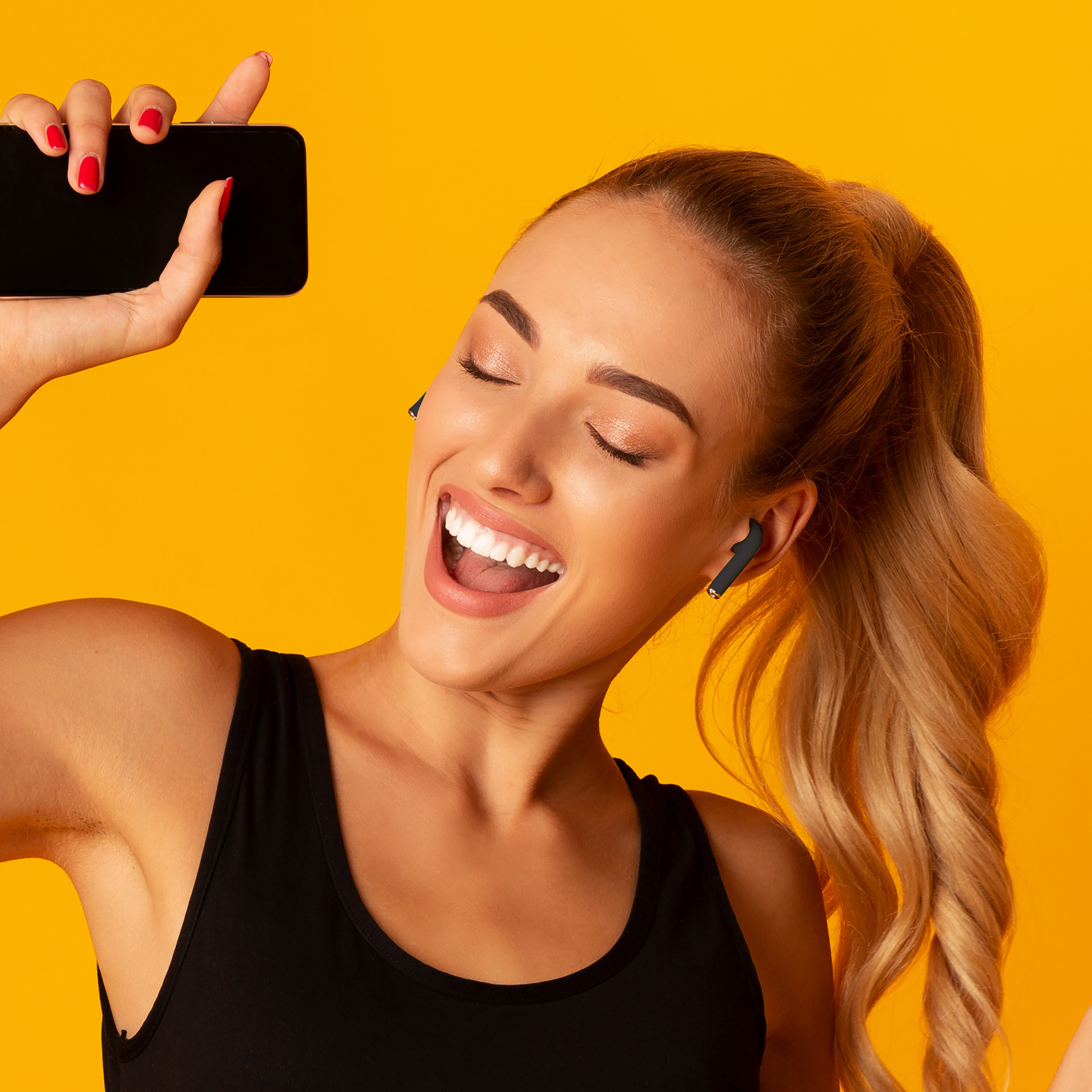 Donna con coda di cavallo, indossa auricolari, tiene il telefono, canta a bocca aperta. Sfondo giallo.