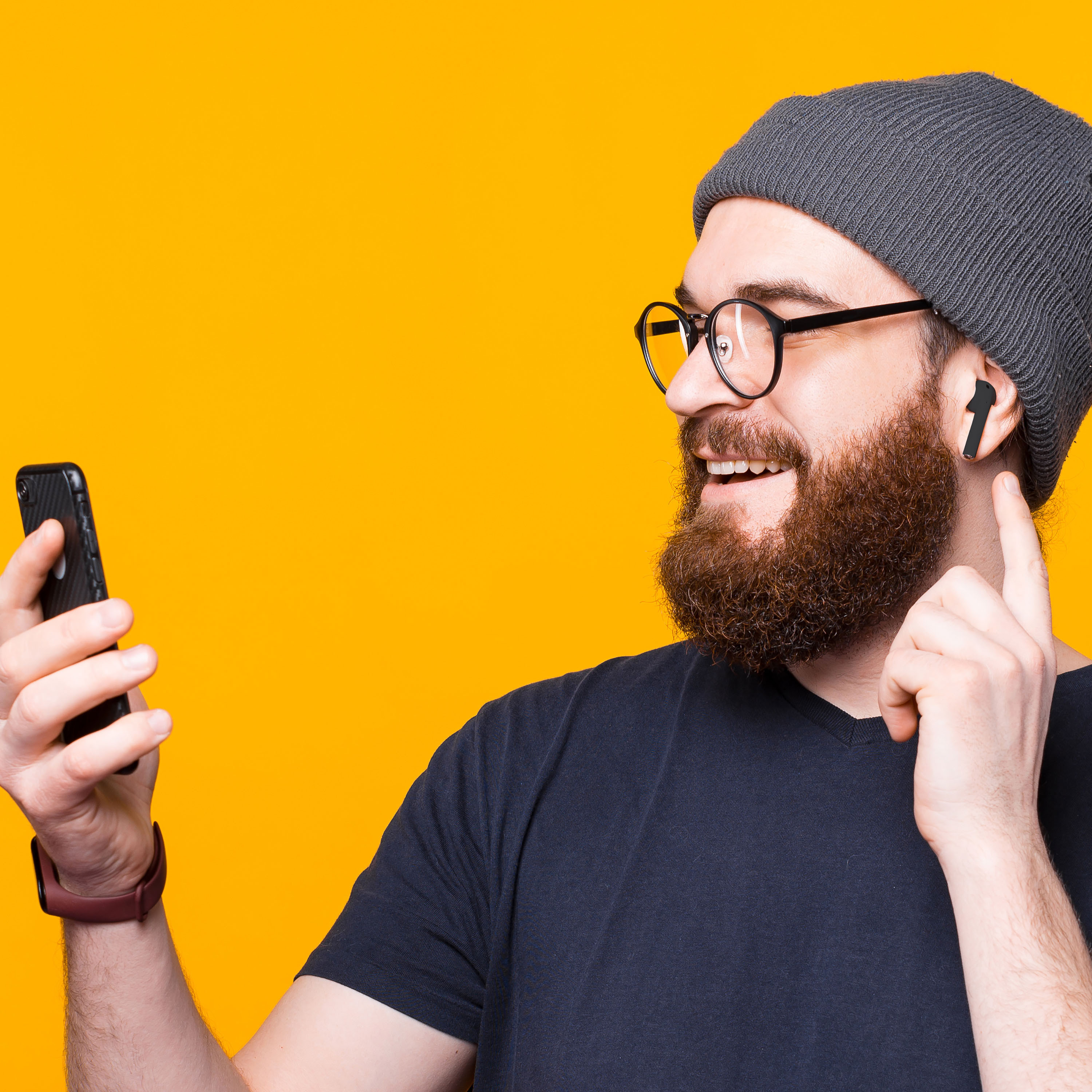 Un uomo con barba e occhiali tiene in mano un telefono e indica un auricolare nell'orecchio.
