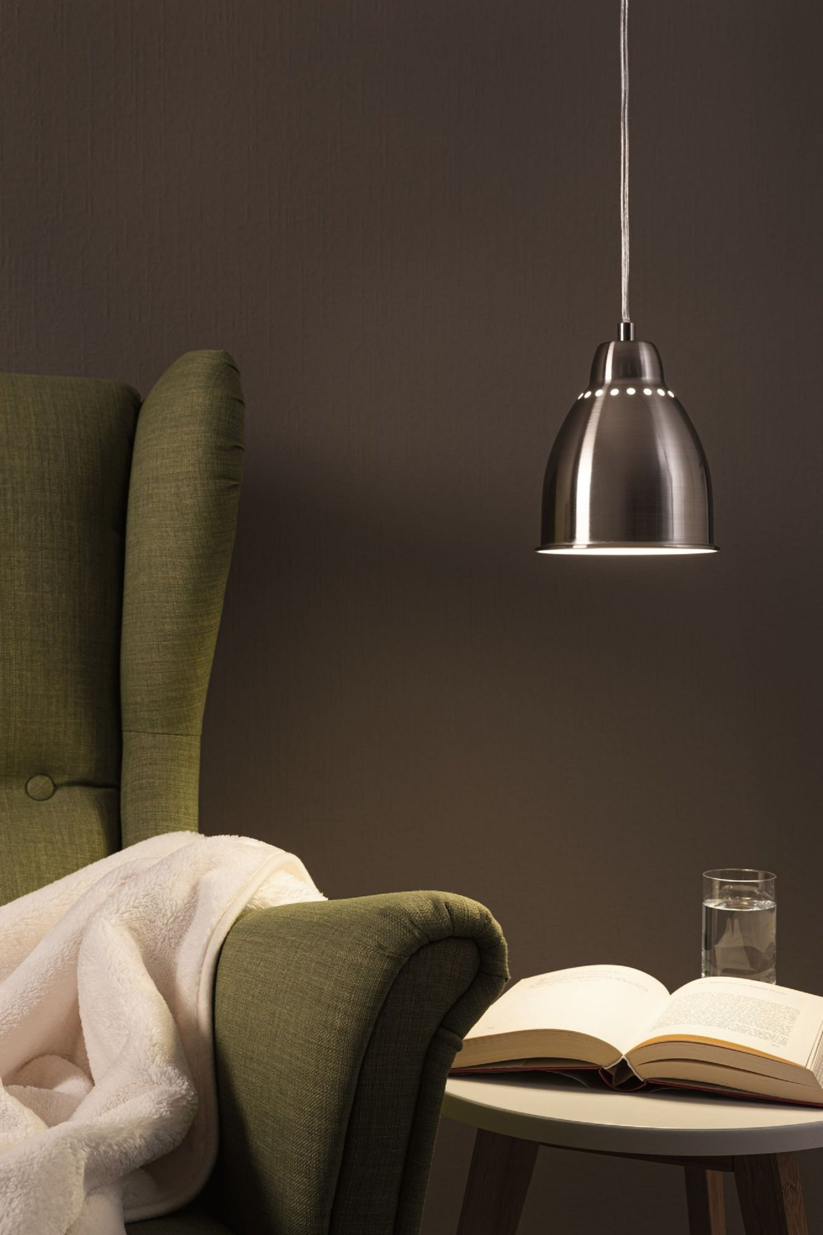 Una lampada a sospensione è appesa sopra una poltrona verde con una coperta. Un libro e un bicchiere sul tavolino.