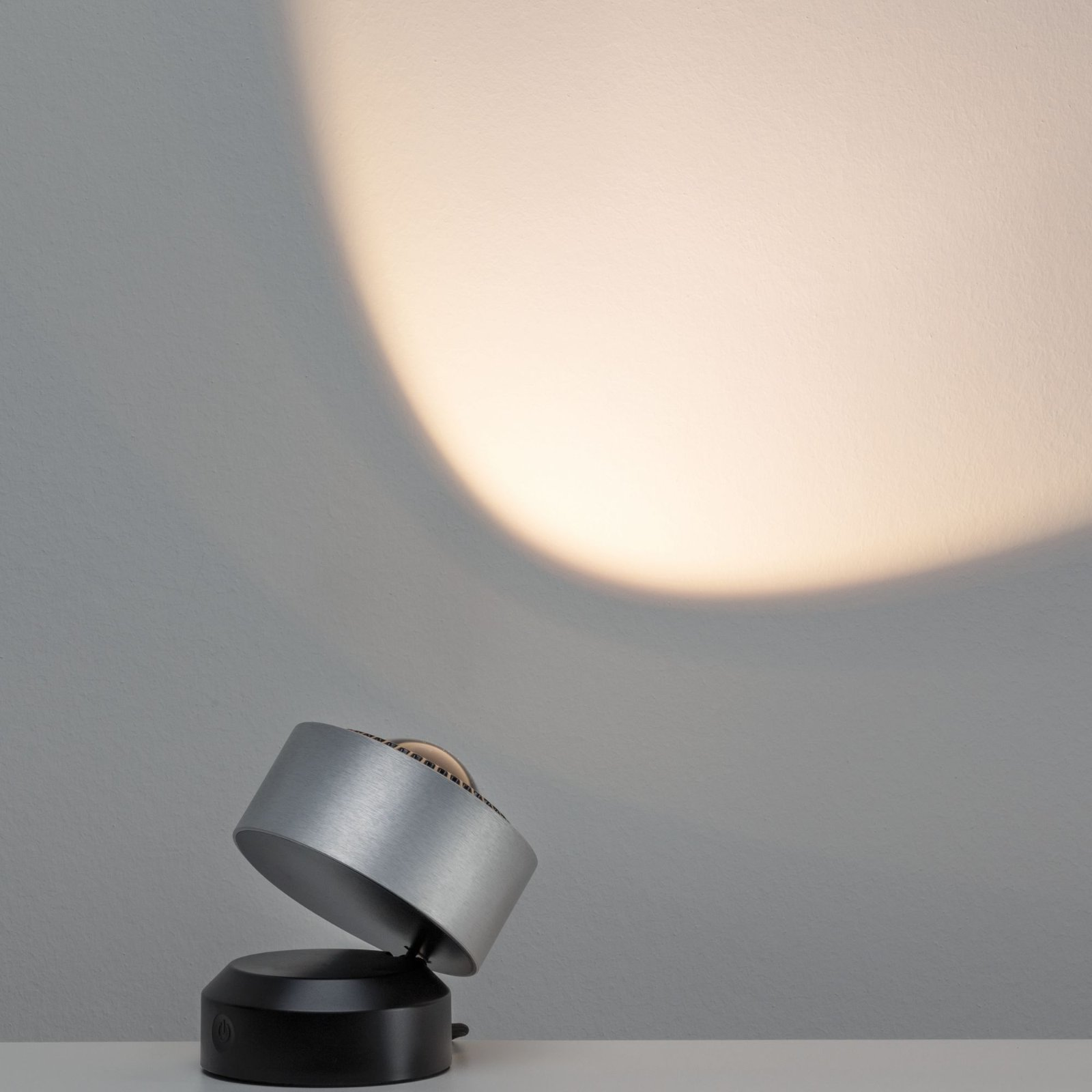 Ein Spot auf einer weißen Oberfläche. Das Licht ist Metall und schwarz. Das Licht scheint auf eine graue Wand.