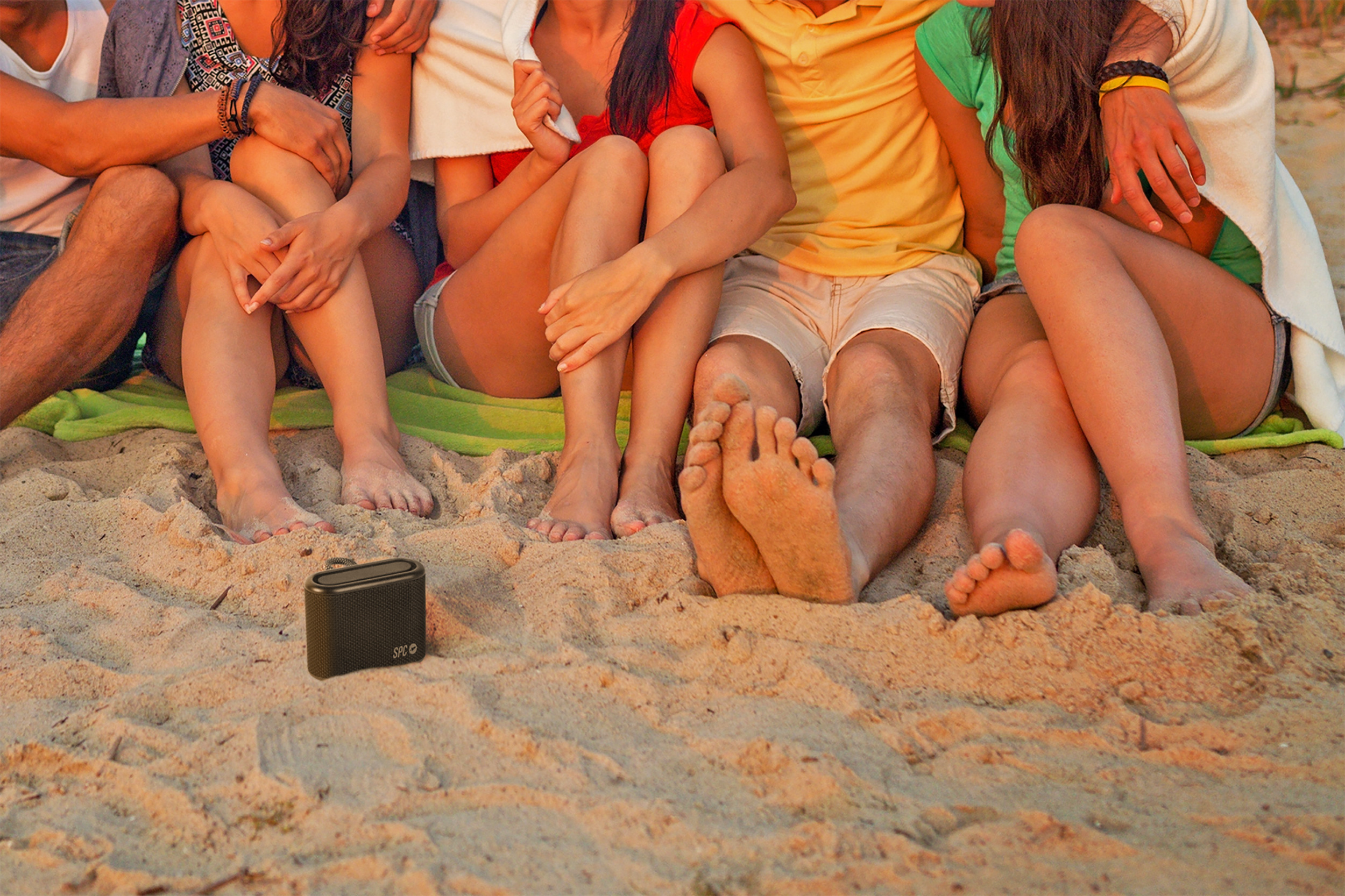 Grupo de personas sentadas en la playa, con un pequeño altavoz en primer plano, pies visibles.
