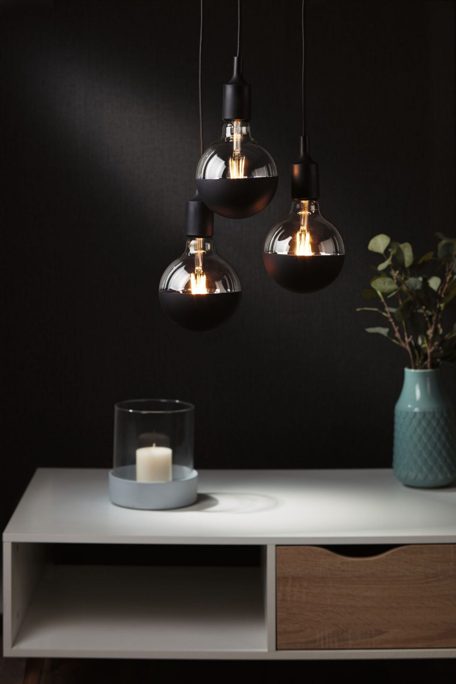 Trzy wiszące żarówki z czarnymi górnymi częściami i biała półka z dekoracjami i świecą.