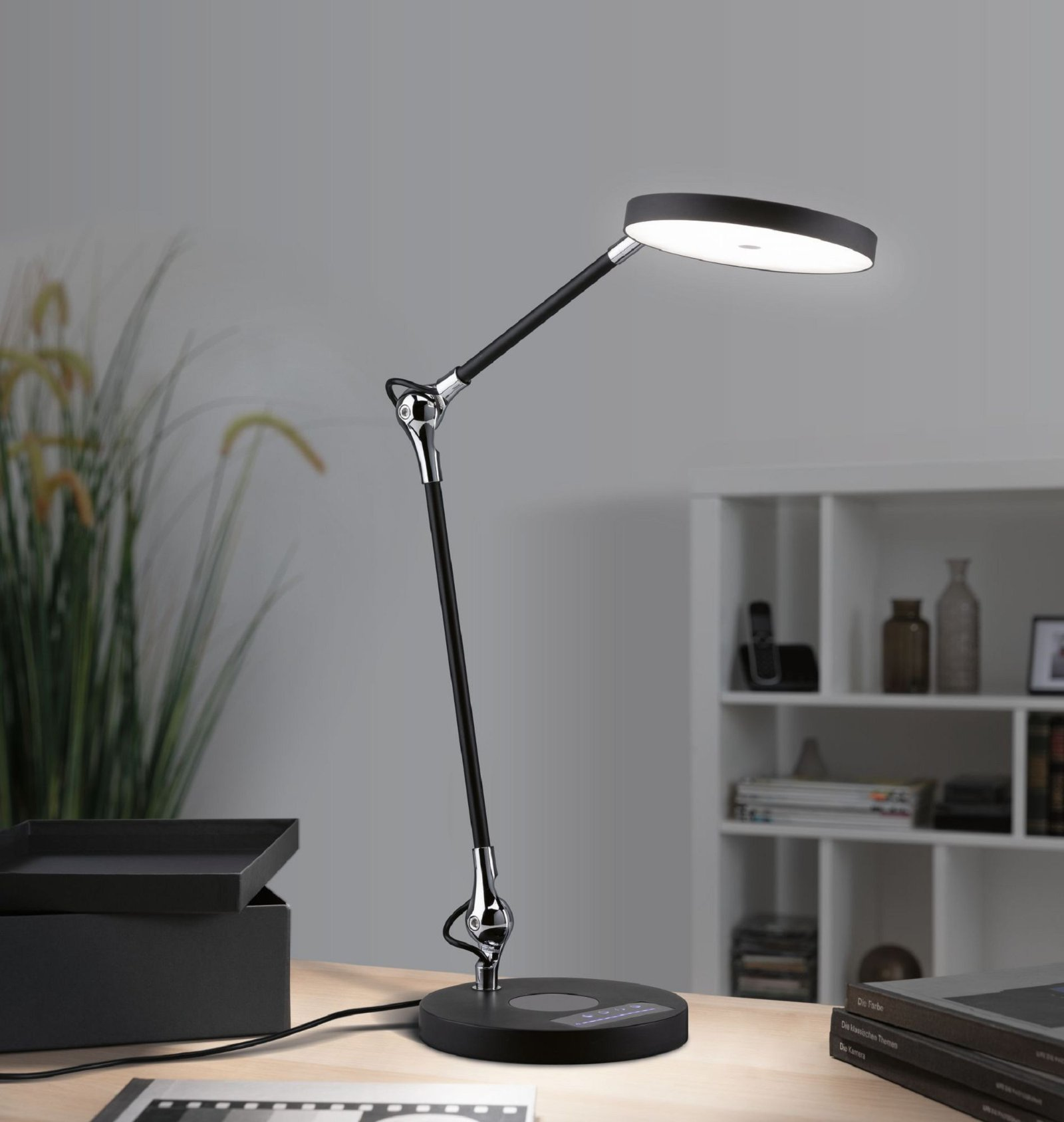 Schwarze und silberfarbene Schreibtischlampe mit verstellbarem Arm, auf einem Holzschreibtisch. Im Hintergrund ein Regal.