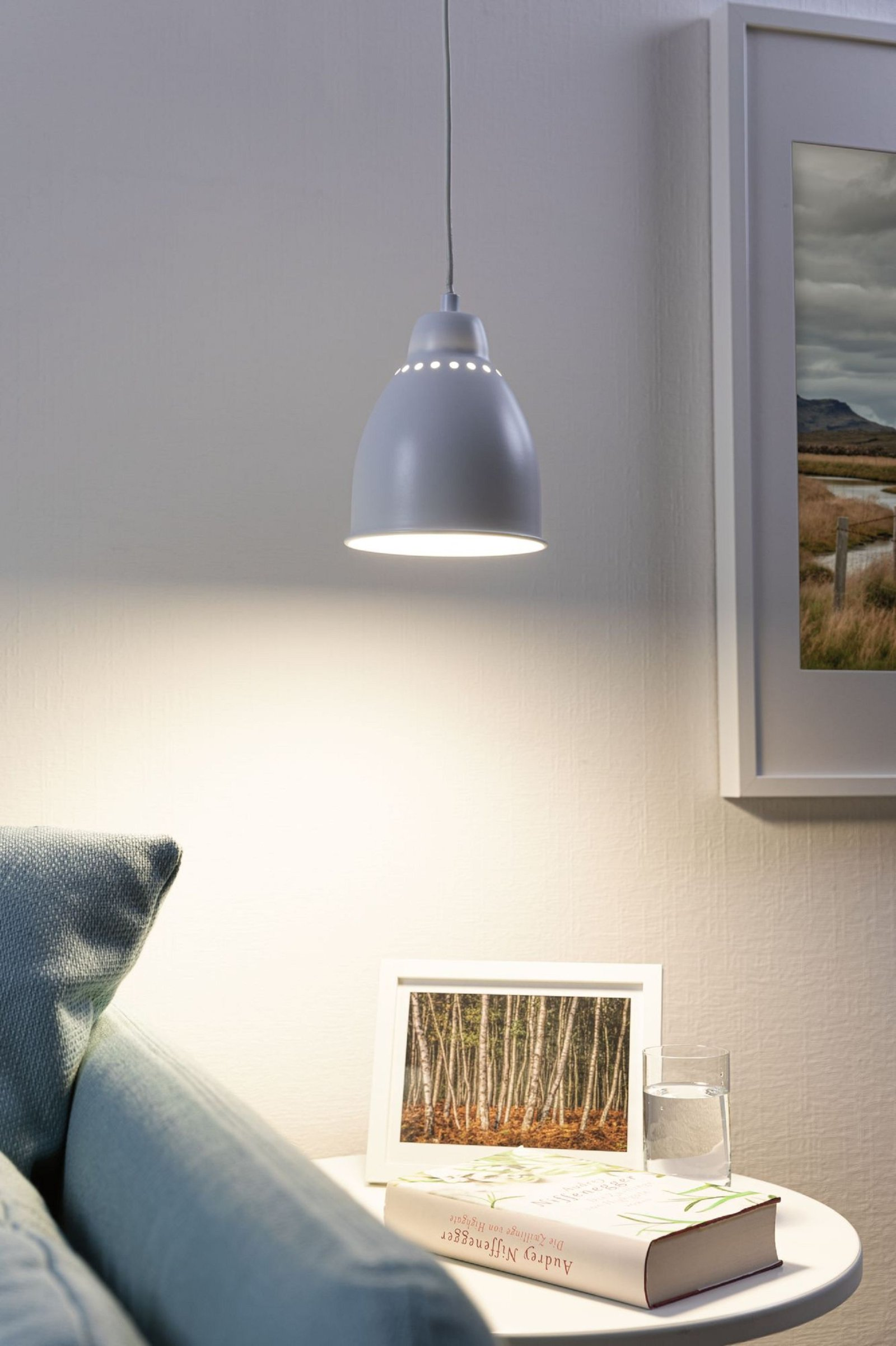 Una lampada a sospensione grigia è appesa sopra un tavolo con un libro, un bicchiere d'acqua e una cornice.