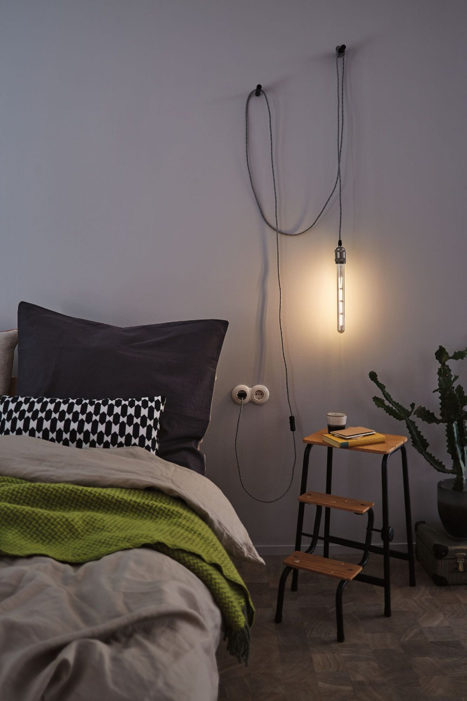 Szene am Bett mit einer hängenden Glühbirne. Es gibt einen hölzernen Tritthocker und eine Pflanze. Graue und weiße Wände.