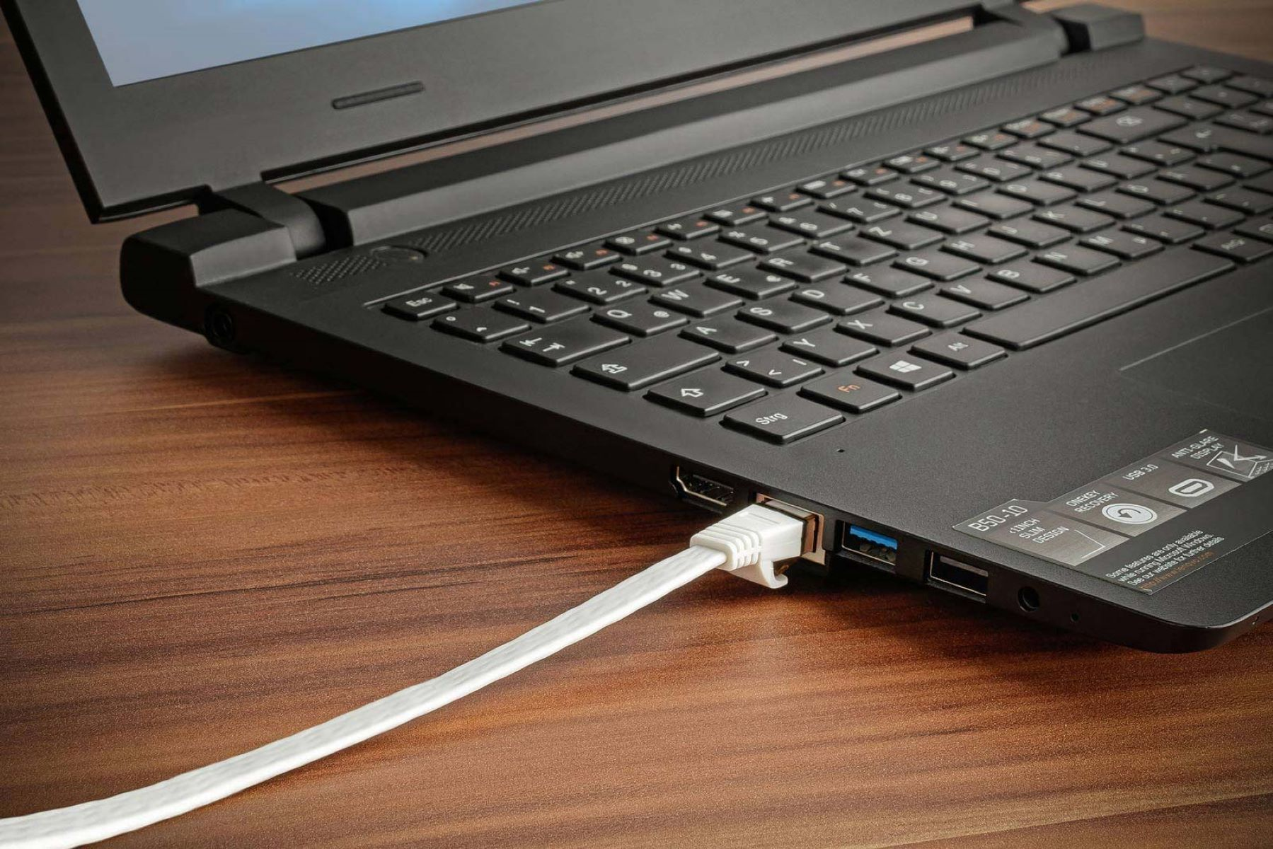 Ein schwarzer Laptop mit einem weißen Kabel, das eingesteckt ist, liegt auf einem Holztisch.