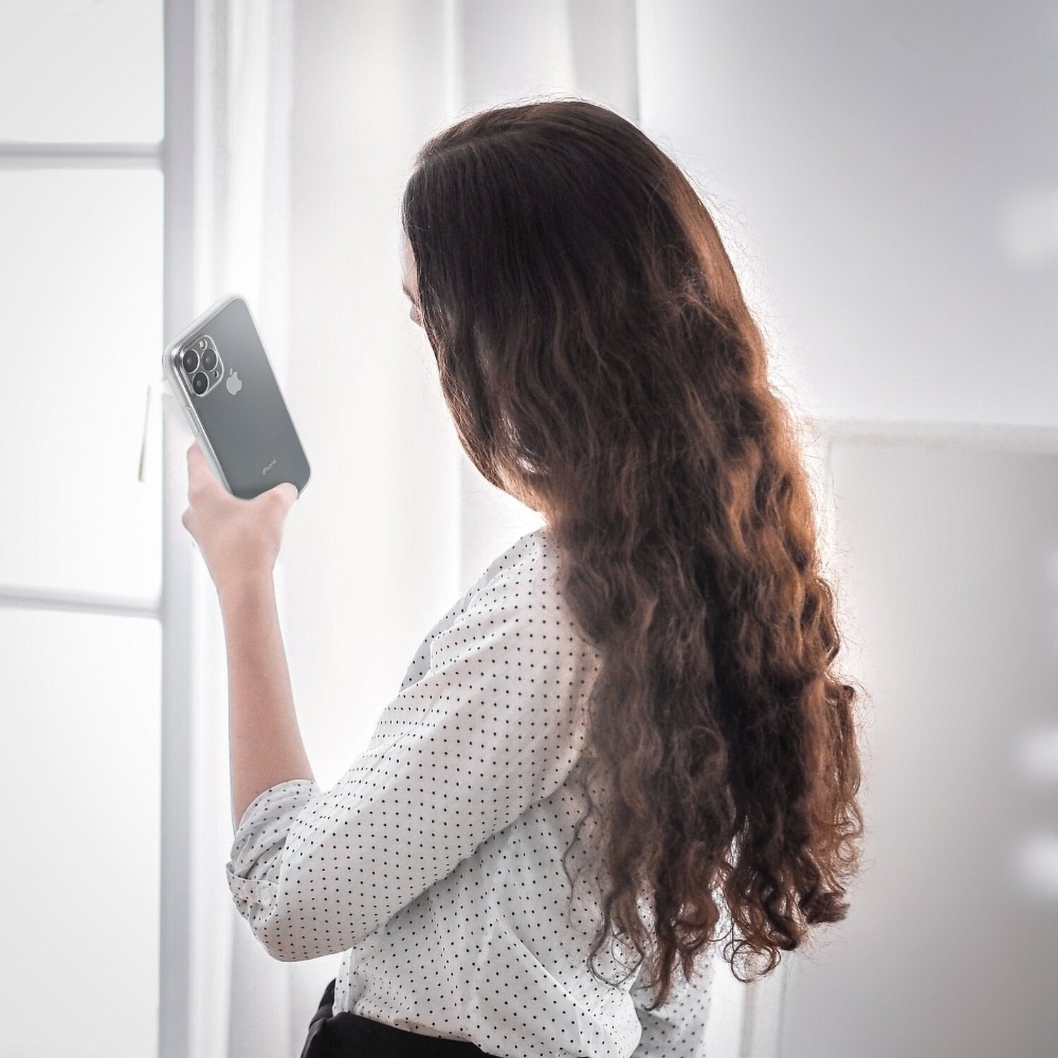 Frau mit langen, welligen Haaren hält ein Telefon und blickt auf den Bildschirm, nahe einem Fenster.