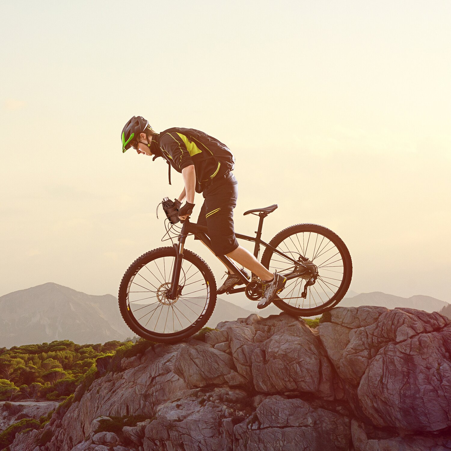 Ein Biker fährt mit einem Mountainbike auf felsigem Gelände. Der Himmel ist hell. Der Hintergrund zeigt Berge.