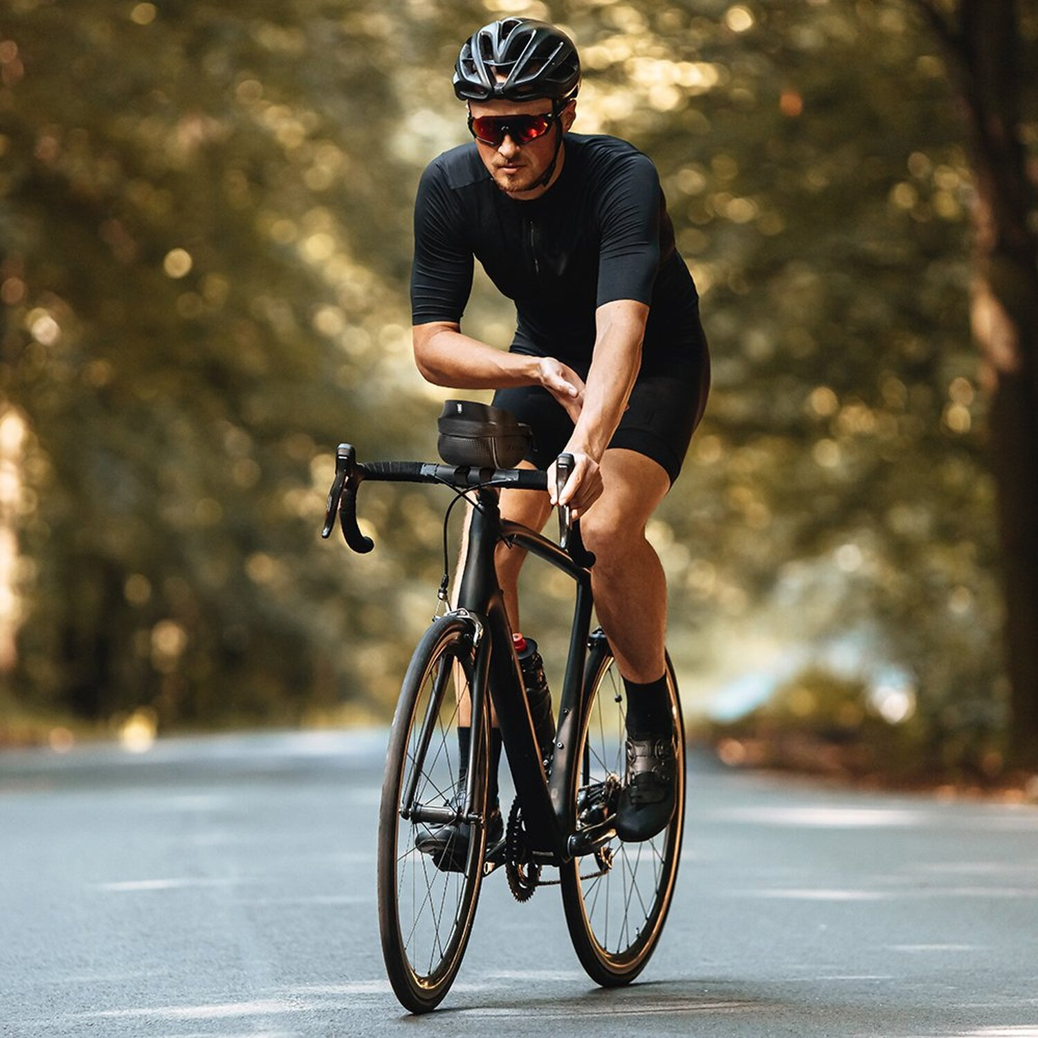 Radfahrer in schwarzer Kleidung fährt auf einer schwarzen Rennrad auf einer Straße durch Wald.