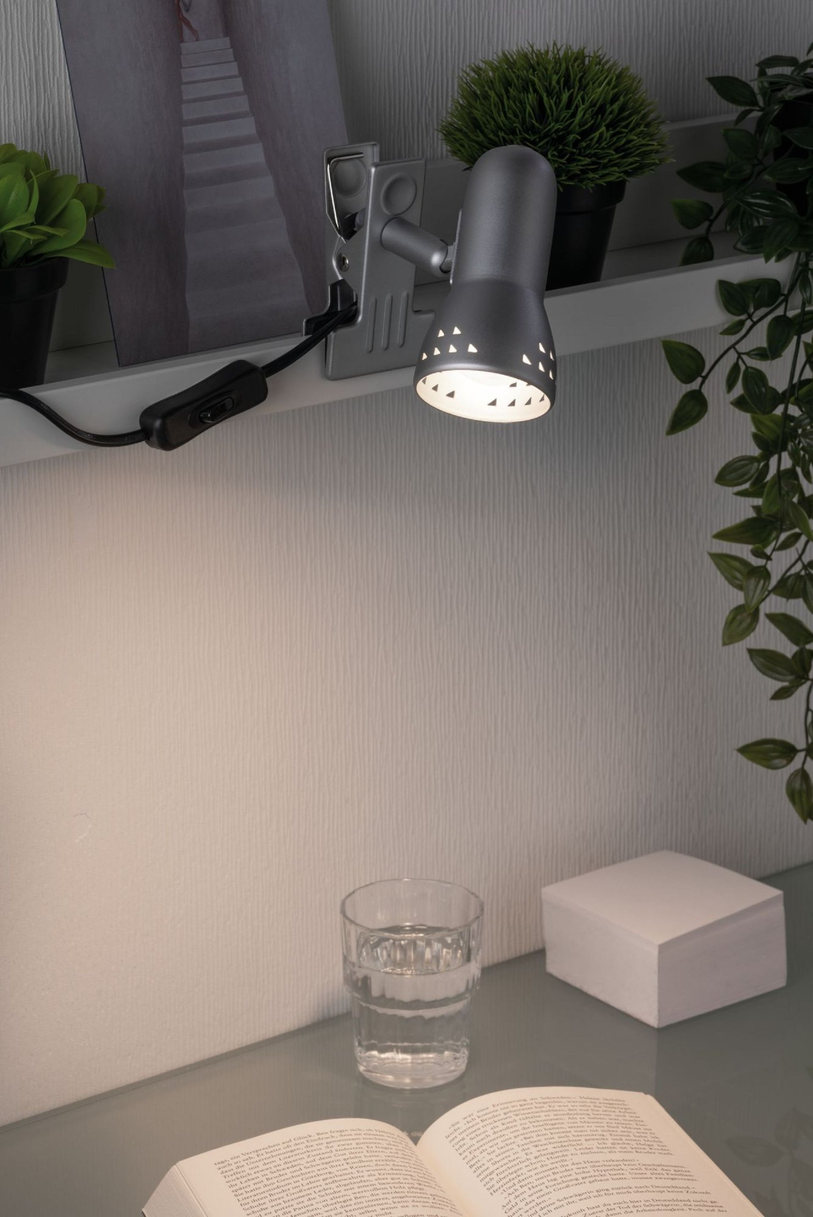 Srebrna lampa zaciskowa na półce, oświetlająca białą ścianę. Szklanka wody i otwarta książka z przodu.