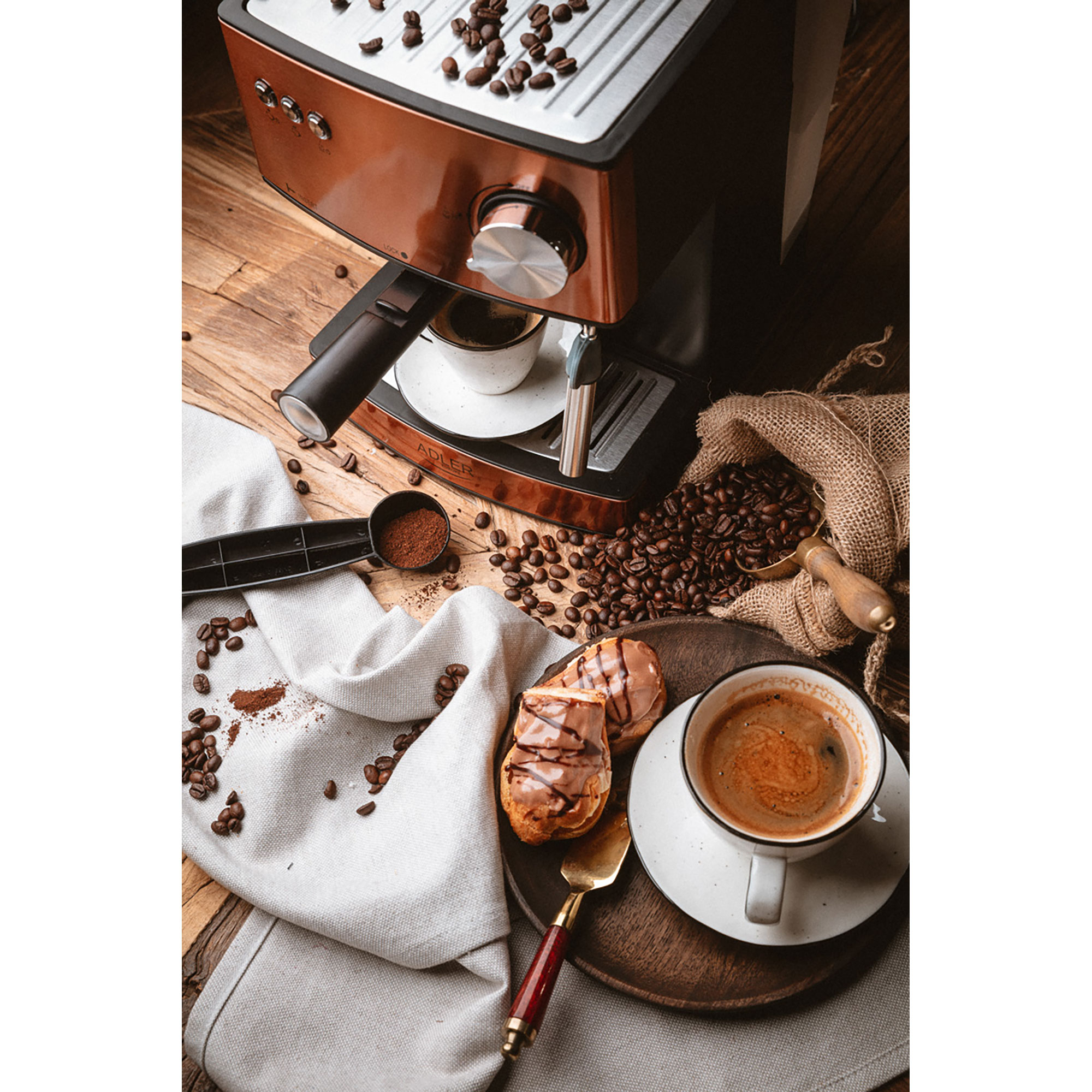 Macchina da caffè con caffè, pasticceria, chicchi e cucchiaio su un tavolo di legno con un panno bianco.